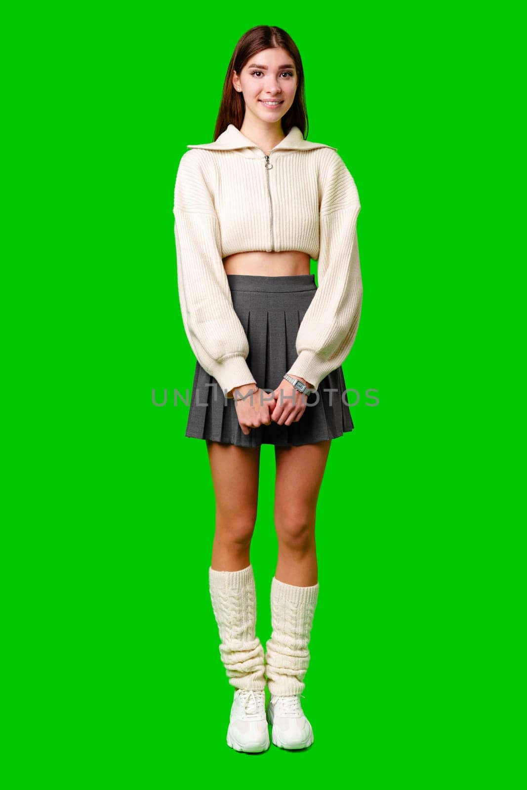 A cheerful young woman stands with confidence against a green backdrop, dressed in a white zippered sweater and a grey skirt. Her hands are gently clasped in front of her as she conveys a friendly demeanor with her smile.