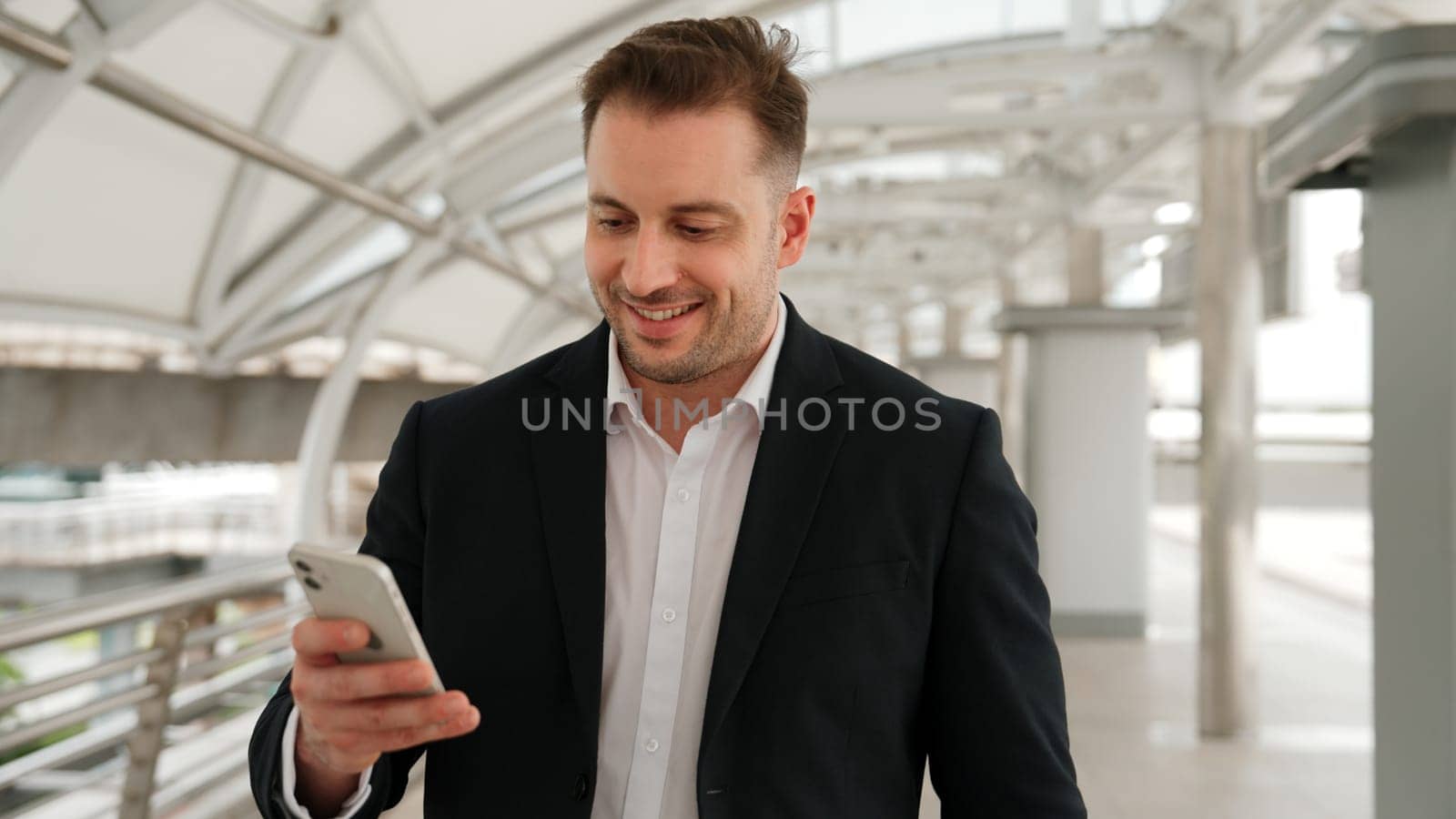 Skilled businessman looking or checking his mobile phone while walking at bridge for going to workplace. Happy manager using telephone to send massage to marketing team and receive good news. Urbane.