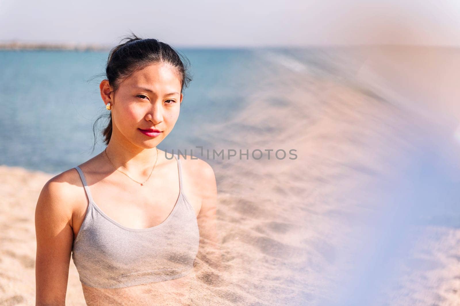 young contemplative asian female in sportswear at beach looking at camera, concept of beauty and purity, flare effect