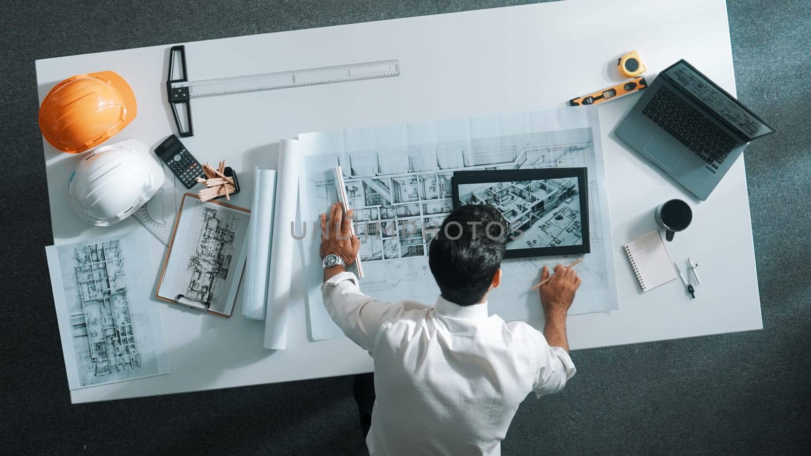Top view of skilled architect looking at project plan at tablet. Aerial view of civil engineer planning while checking building blueprint at meeting room with safety helmet and laptop. Alimentation.