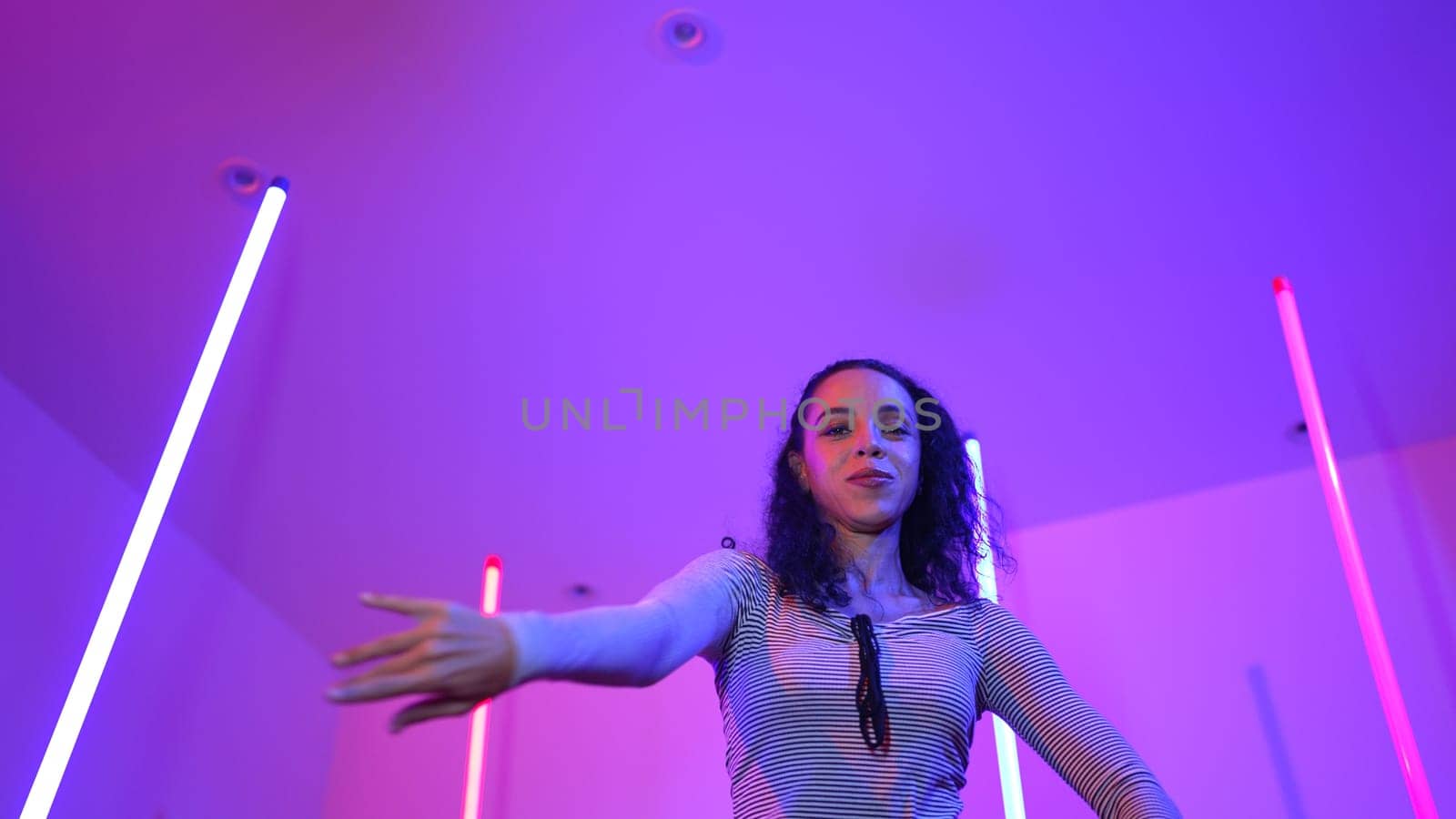 Low angle camera of hispanic dancer looking at camera and dancing to city pop music. Professional attractive hipster moving to hip hop song with purple led or neon light. Stylish cloth. Regalement.