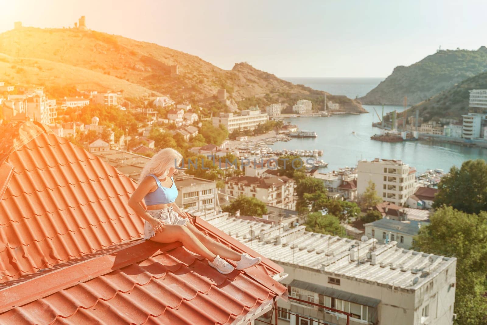 Woman sits on rooftop, enjoys town view and sea mountains. Peaceful rooftop relaxation. Below her, there is a town with several boats visible in the water. Rooftop vantage point