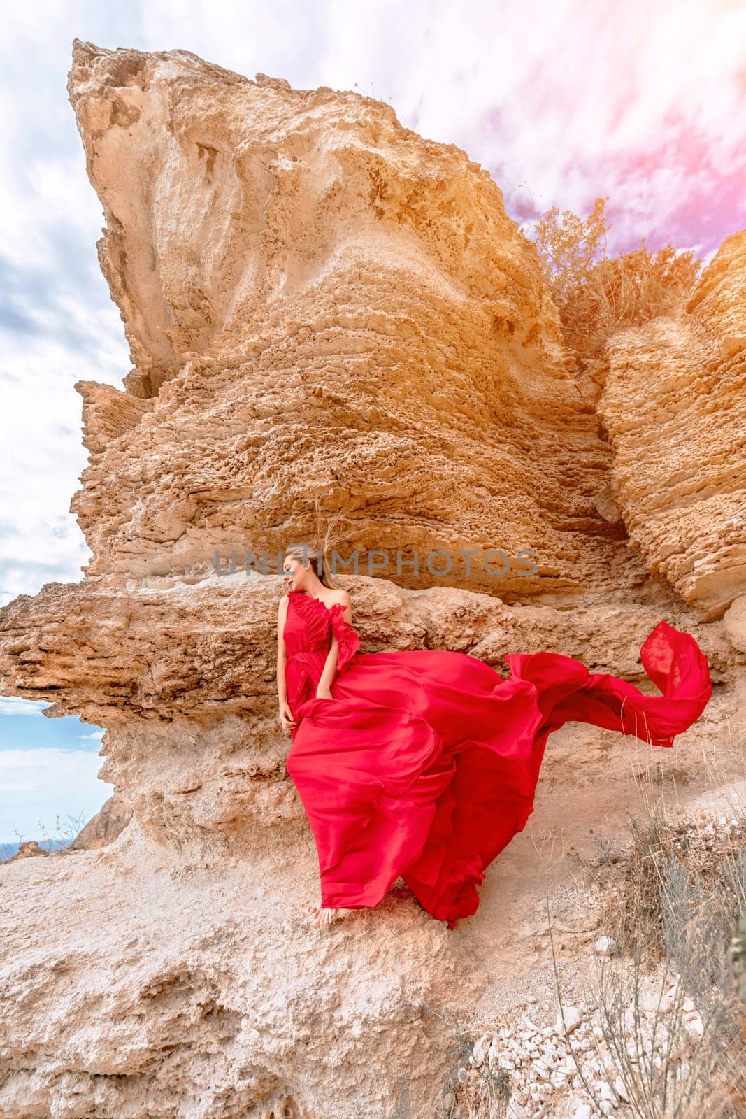 woman red silk dress stands by the ocean, with mountains in the background, as her dress sways in the breeze
