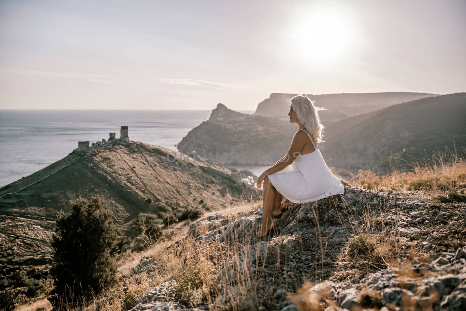 A woman is sitting on a hillside overlooking the ocean. She is wearing a white dress and has blonde hair. The scene is serene and peaceful, with the ocean in the background. by Matiunina