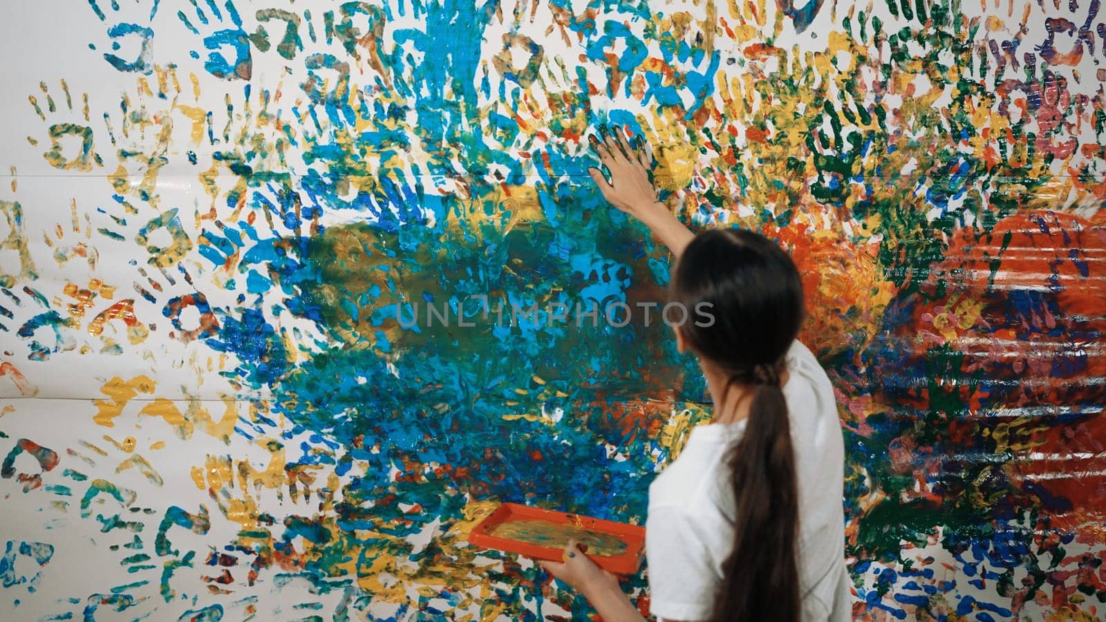 Young happy girl holding the color tray and paint colorful stained wall to express idea and enhance imagination with hand print. Caucasian girl stamp hand to decorate room in art lesson. Edification.