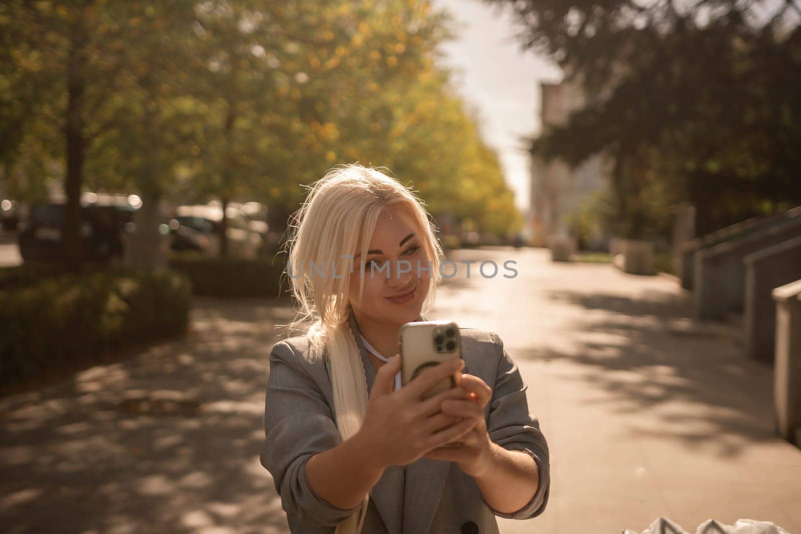 A woman is taking a picture of herself with her cell phone. She is wearing a gray jacket and scarf. The scene is set in a city with trees and cars in the background. by Matiunina