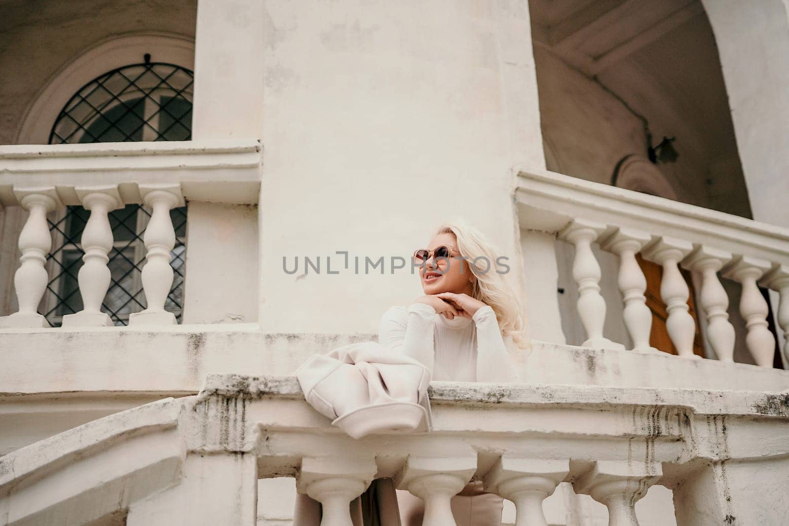 A woman is sitting on a white railing, looking out at the street. She is wearing sunglasses and a white shirt. The scene is set in front of a building with white pillars