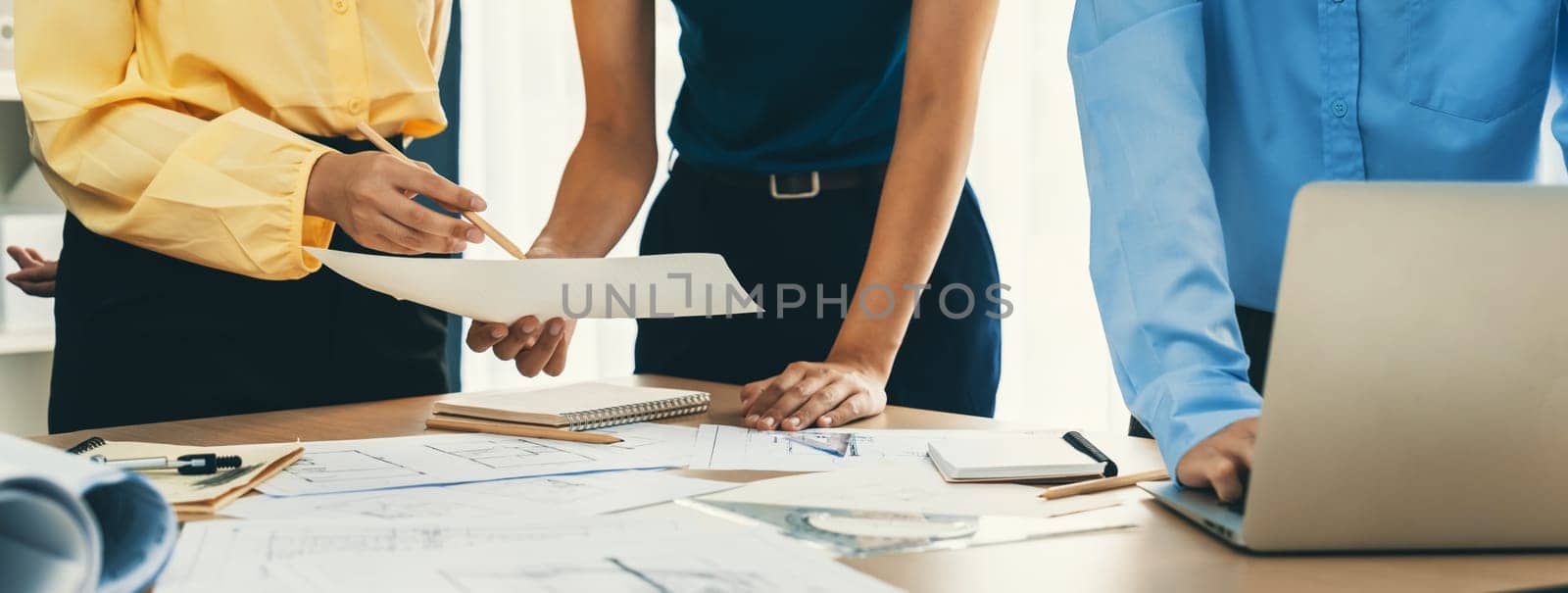Architect team discuss about blueprint design at modern meeting room while her coworker working on laptop on table with blueprint and architectural equipment scattered around. Closeup. Delineation.