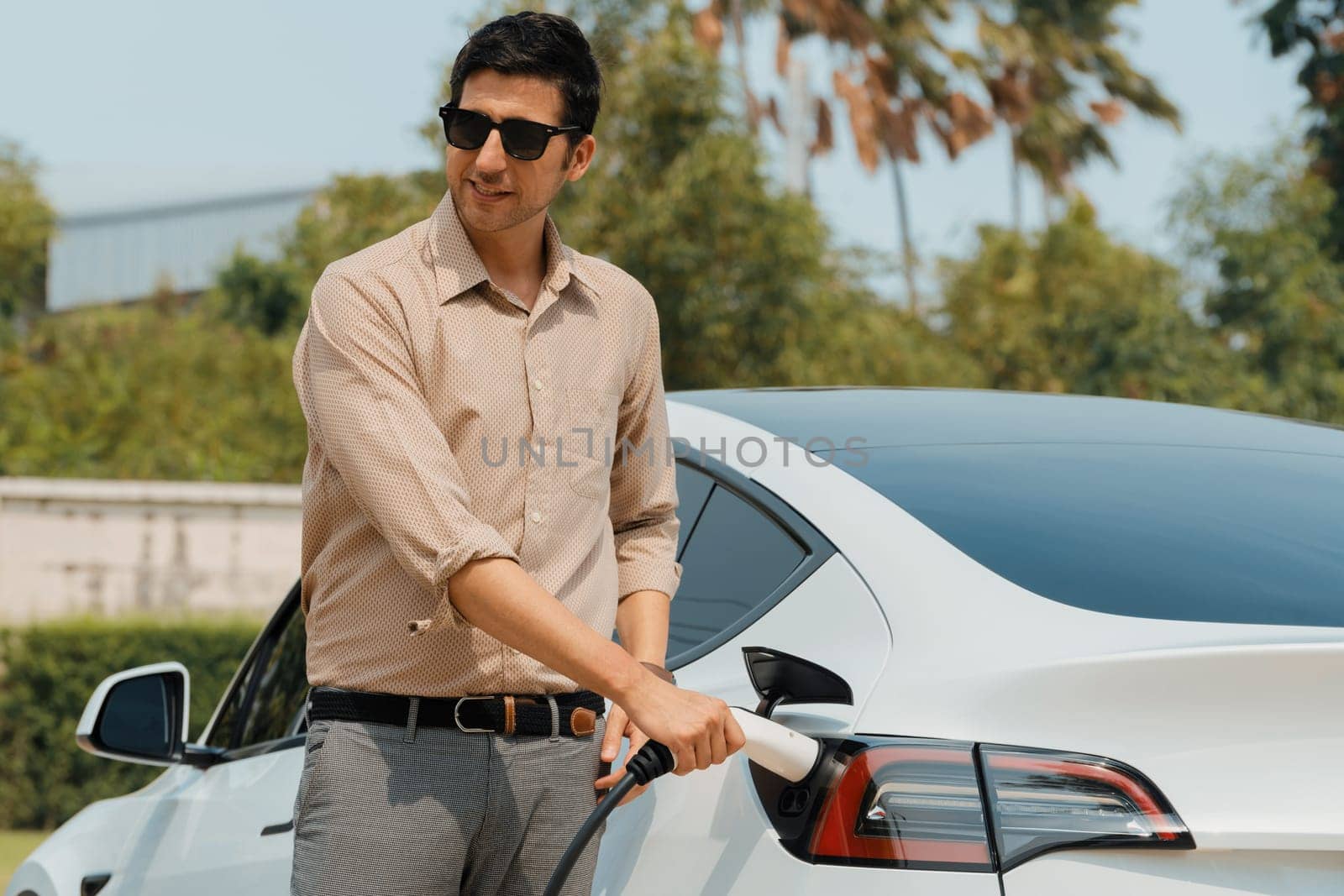 Young man recharge electric car's battery from charging station in outdoor green city park in springtime. Rechargeable EV car for sustainable environmental friendly urban travel lifestyle. Expedient