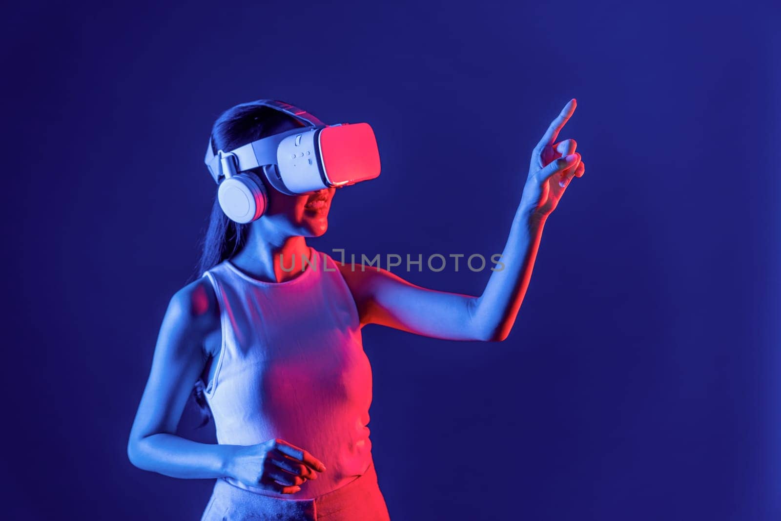Smart female stand with surrounded by cyberpunk neon light wear VR headset connecting metaverse, futuristic cyberspace community technology. Woman using finger pointing virtual object. Hallucination.