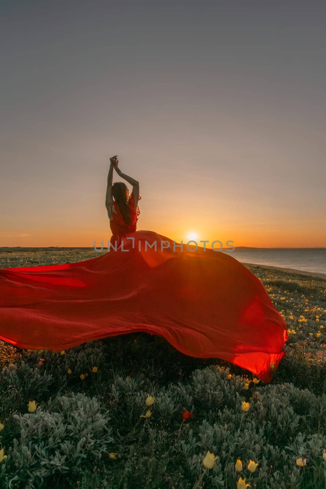A woman in a red dress is standing in a field with the sun setting behind her. She is reaching up with her arms outstretched, as if she is trying to catch the sun. The scene is serene and peaceful. by Matiunina
