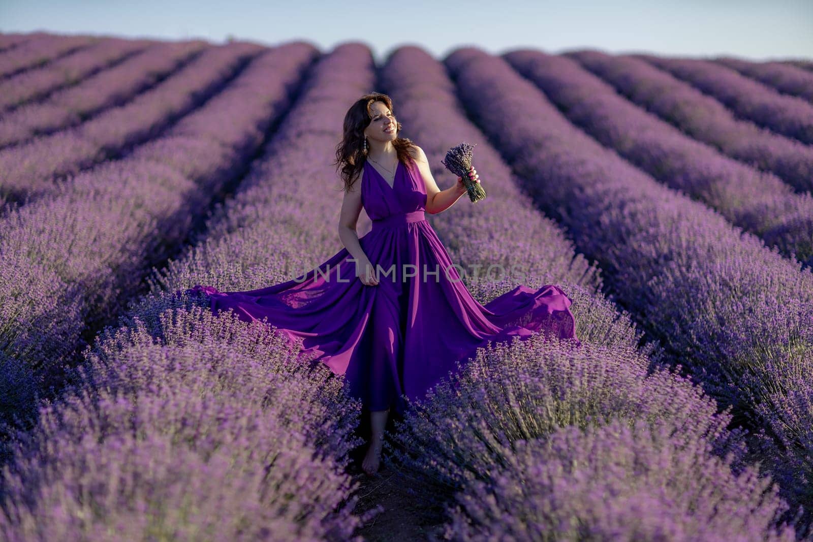 A woman in a purple dress is standing in a field of lavender. She is holding a bouquet of flowers and she is enjoying the beauty of the flowers. Concept of serenity and appreciation for nature. by Matiunina