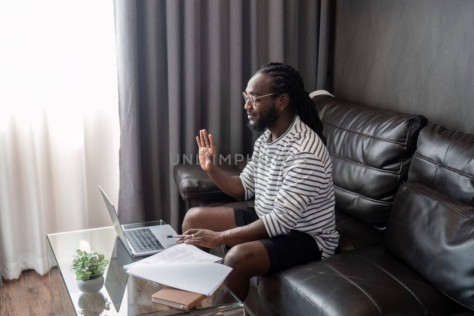 African American man working with laptop computer video calling and conference, remote work while sitting at sofa in living room. Black guy do freelance work at home office by nateemee