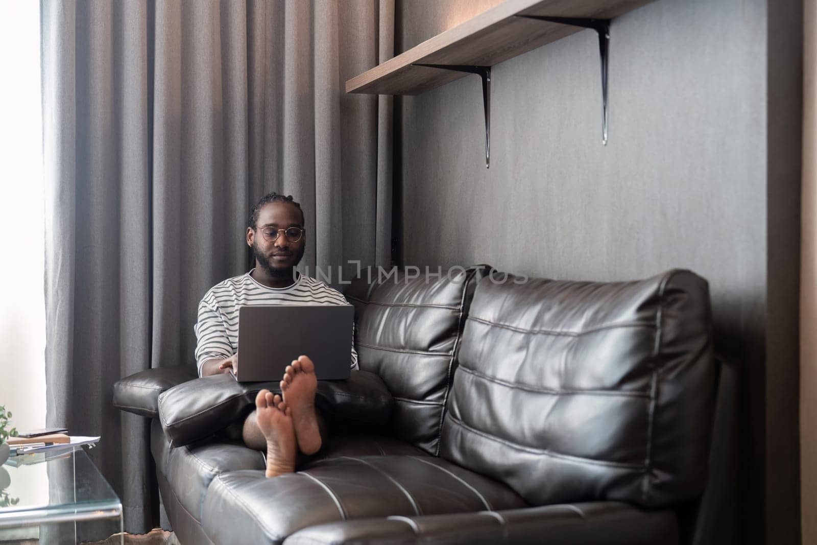 Young African American man holding laptop relaxing sitting on sofa working remote at home.