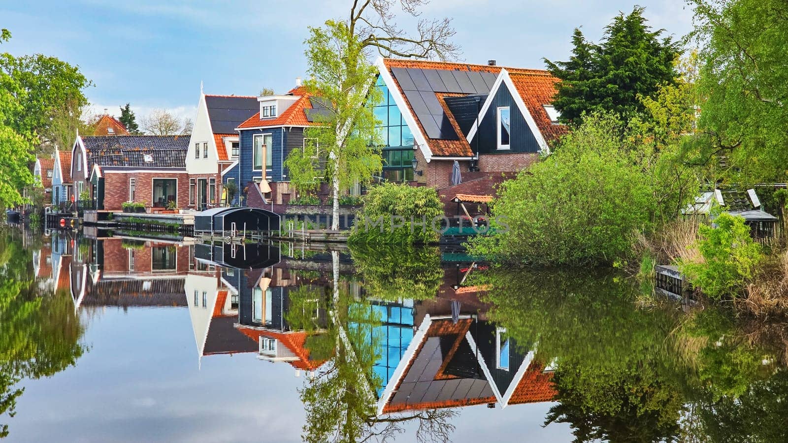 A picturesque scene showing a row of charming houses nestled next to a tranquil body of water, reflecting the clear blue sky and lush greenery by fokkebok