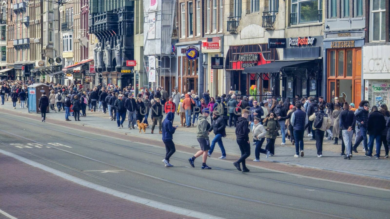 Amsterdam Netherlands 21 April 2024, A diverse crowd of people walks briskly down a bustling city street lined with towering buildings