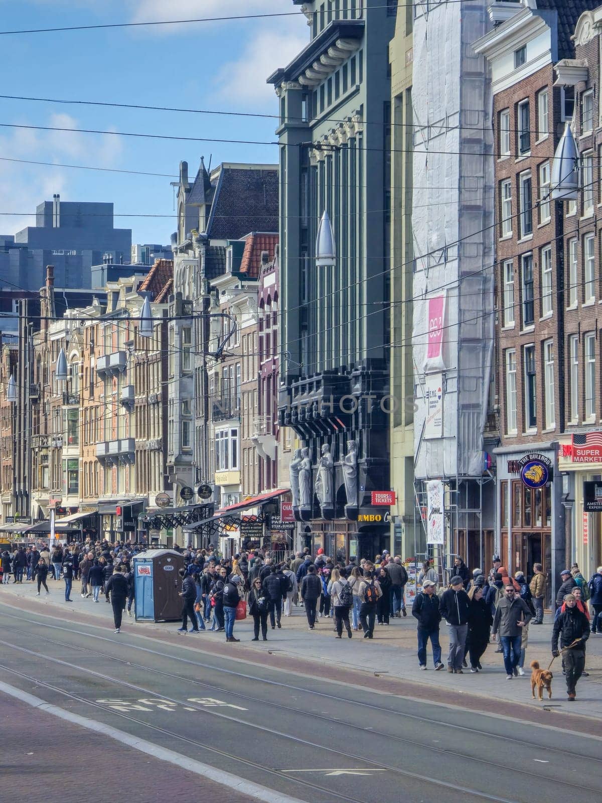 Amsterdam Netherlands 21 April 2024 A diverse group of people walks in a lively hustle down a bustling city street flanked by shops, under a clear blue sky.