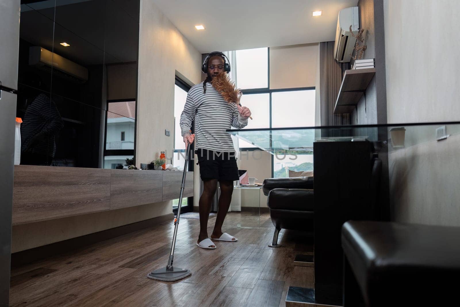 Funny African American is cleaning house doing housework cleaning floor with mop and listening to music in wireless headphones, African American man is dancing and singing by nateemee