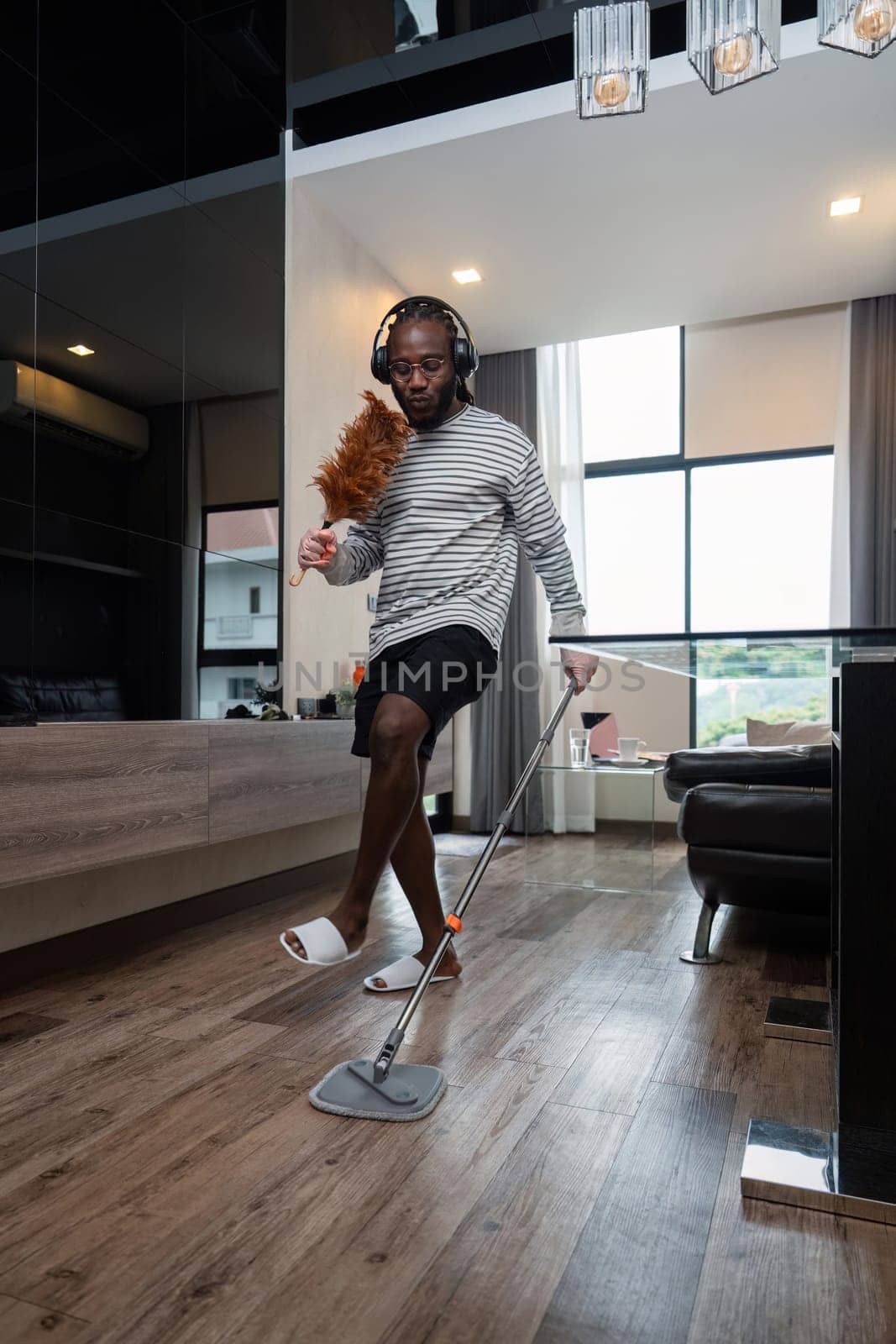 Funny African American is cleaning house doing housework cleaning floor with mop and listening to music in wireless headphones, African American man is dancing and singing.