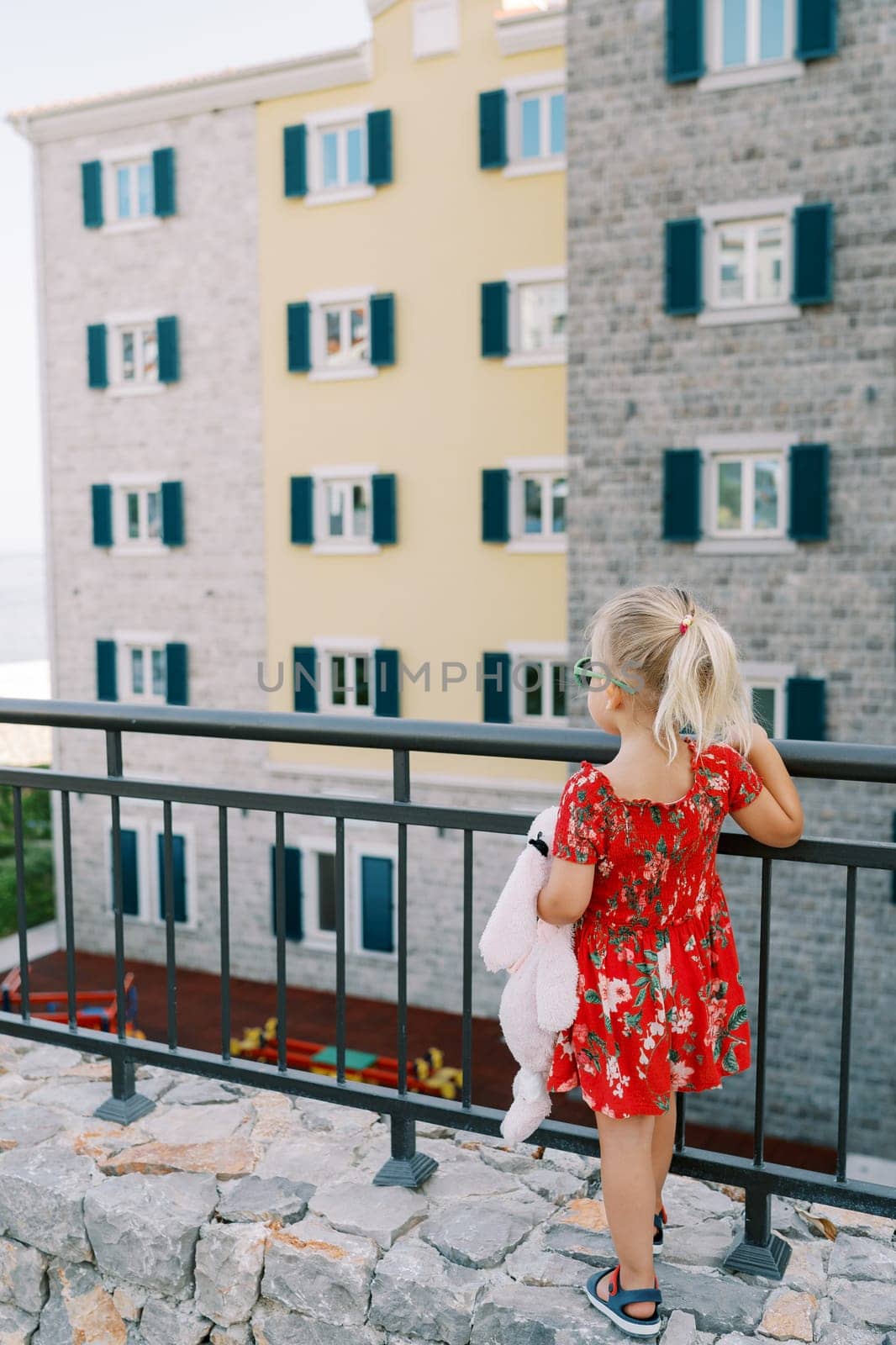 Little girl stands leaning on a fence and looks at an apartment building with a playground. Back view. High quality photo