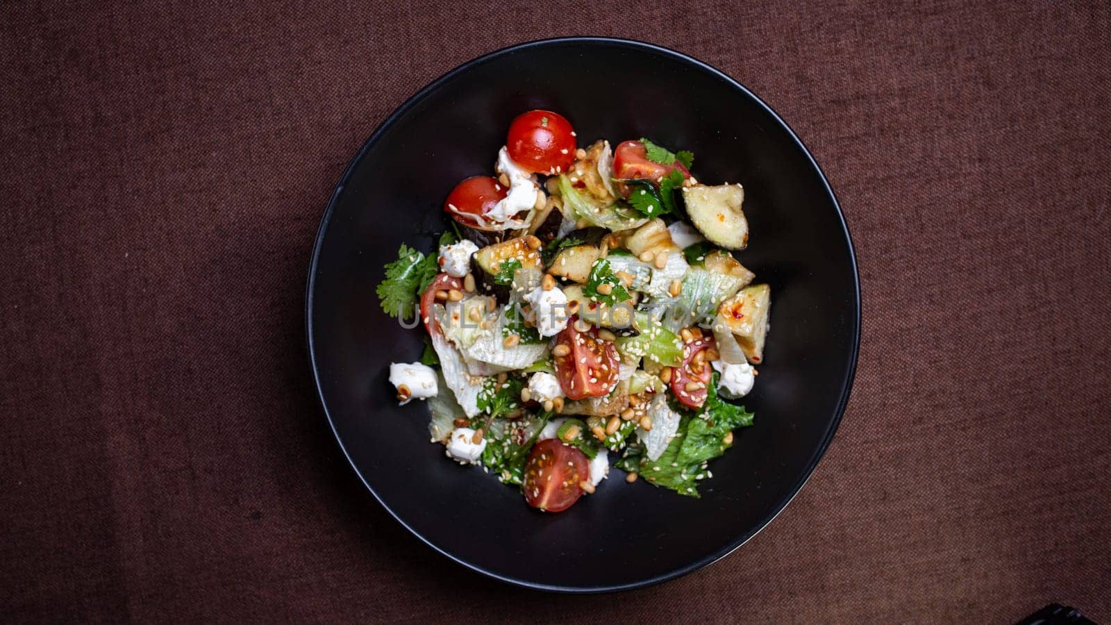 Top view of a delicious black bowl of salad filled with feta, tomatoes, cucumbers, and fresh herbs on a brown table.