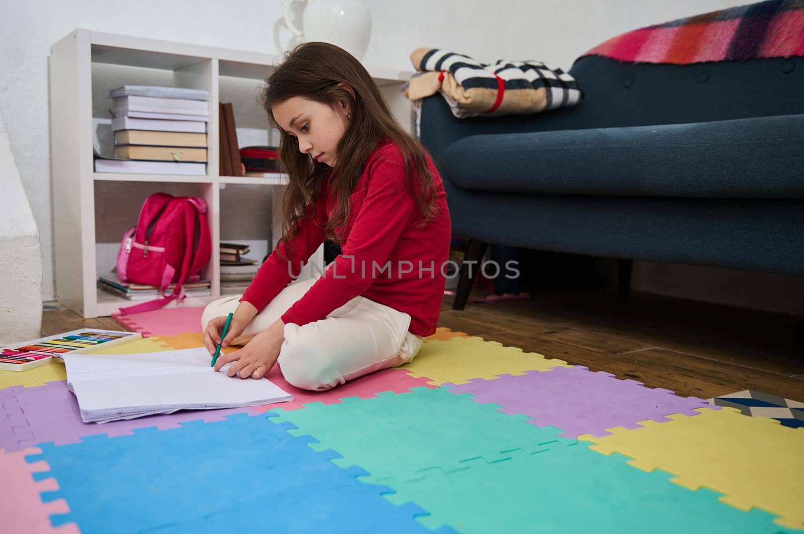 Cute little school girl doing home work in domestic room. Adorable child sitting on a colorful puzzle carpet, writing text. Back to school concept. Children.