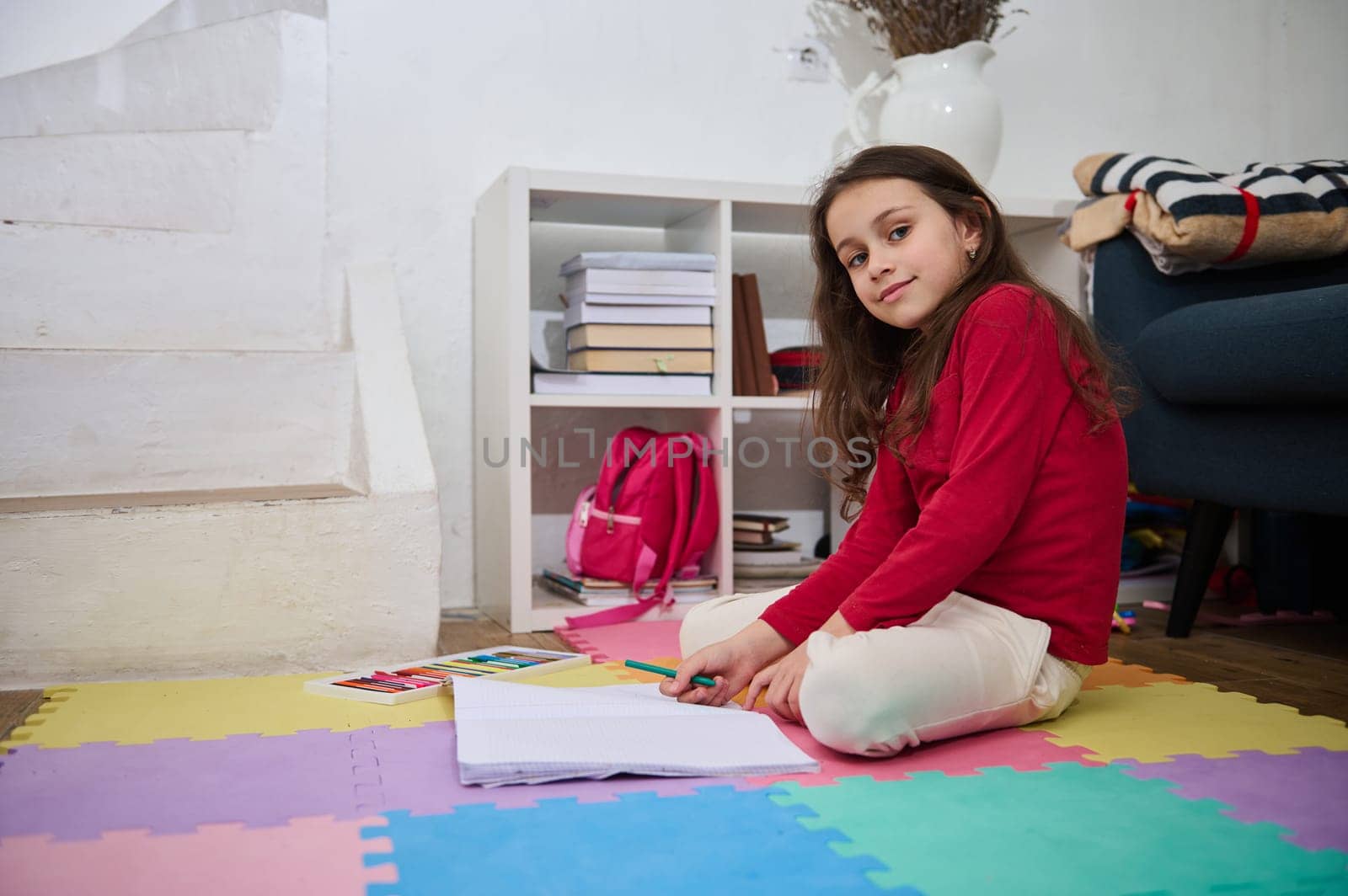 Happy intelligent elementary age school child smiling looking at camera while doing homework at cozy home interior by artgf