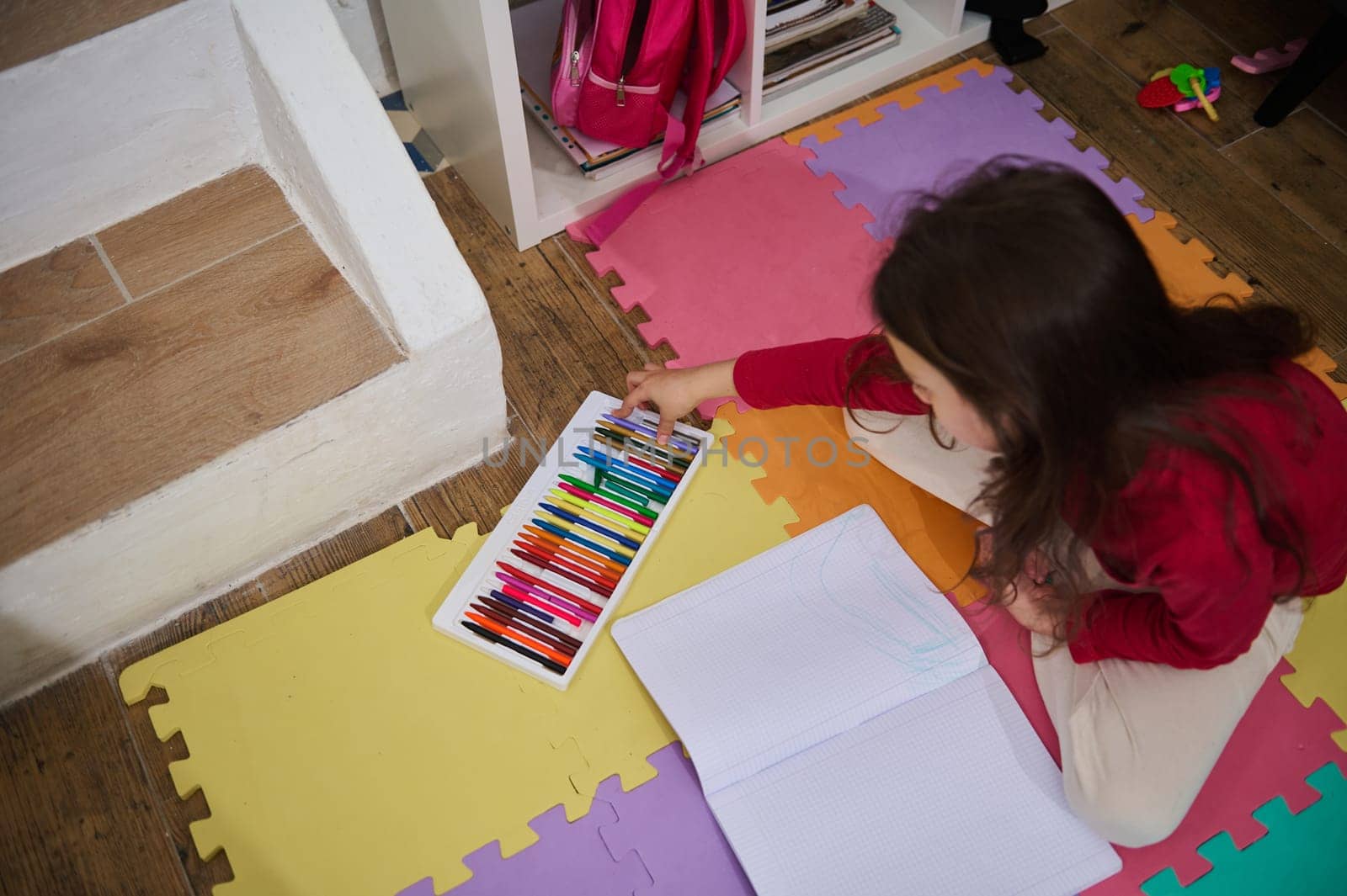 View from above of a little child girl, elementary age school kid taking out colorful pencils from a box, drawing picture at home, lying on a multi colored puzzle pencils. Children. Art and creativity