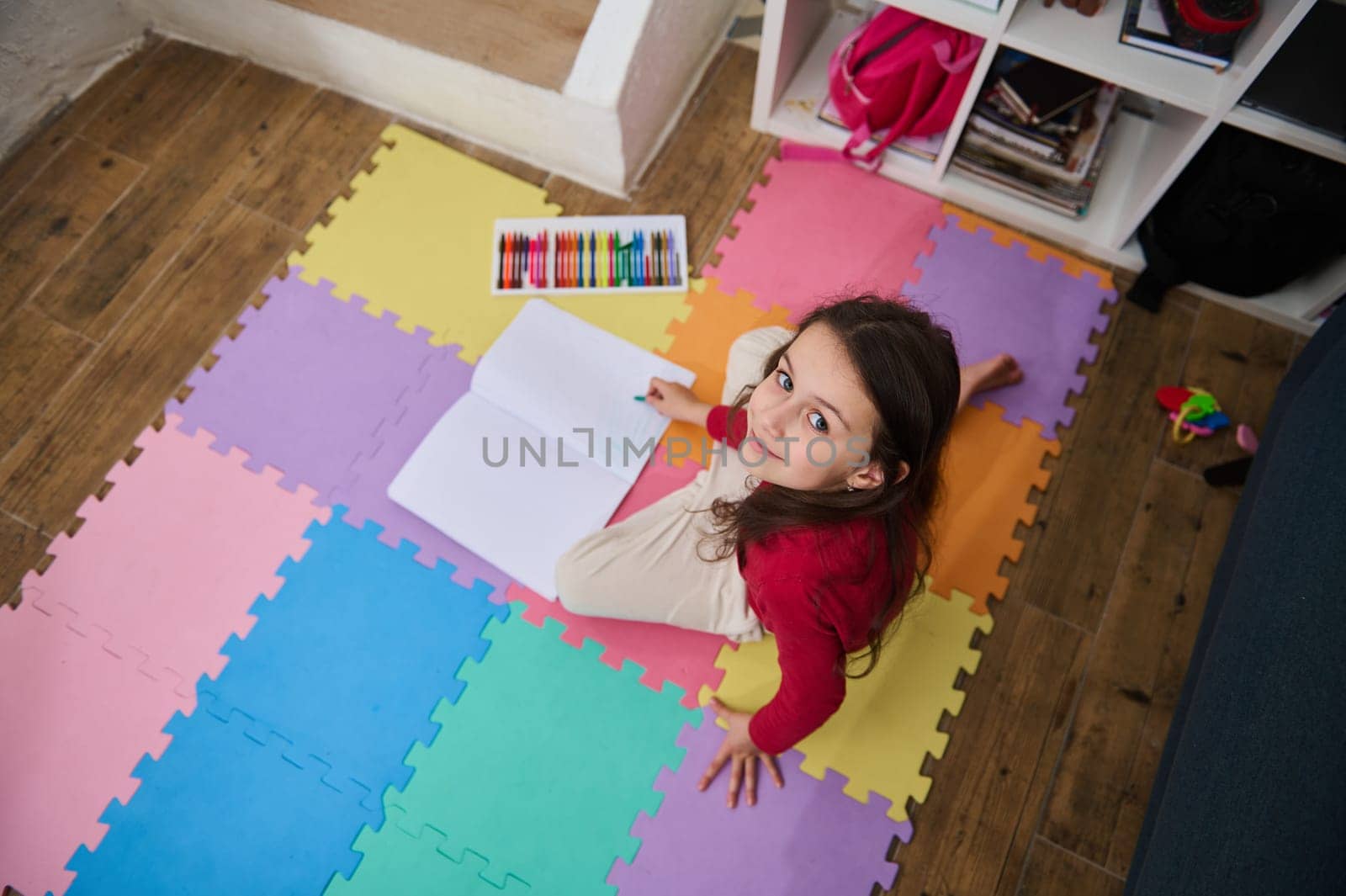 Authentic portrait of beautiful child girl drawing picture using colorful pastel crayons, smiling looking at camera, sitting on a puzzle carpet at home. Art and kids education concept. View from above
