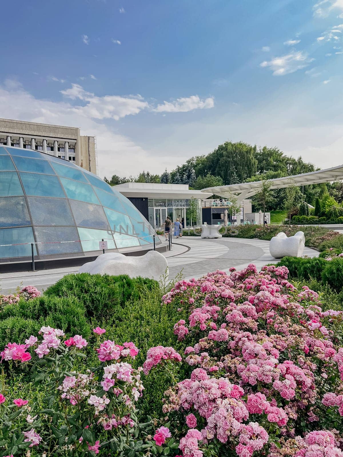 Architectural building with a large glass dome, surrounded by a colorful flower garden. Clear blue sky with fluffy white clouds overhead.