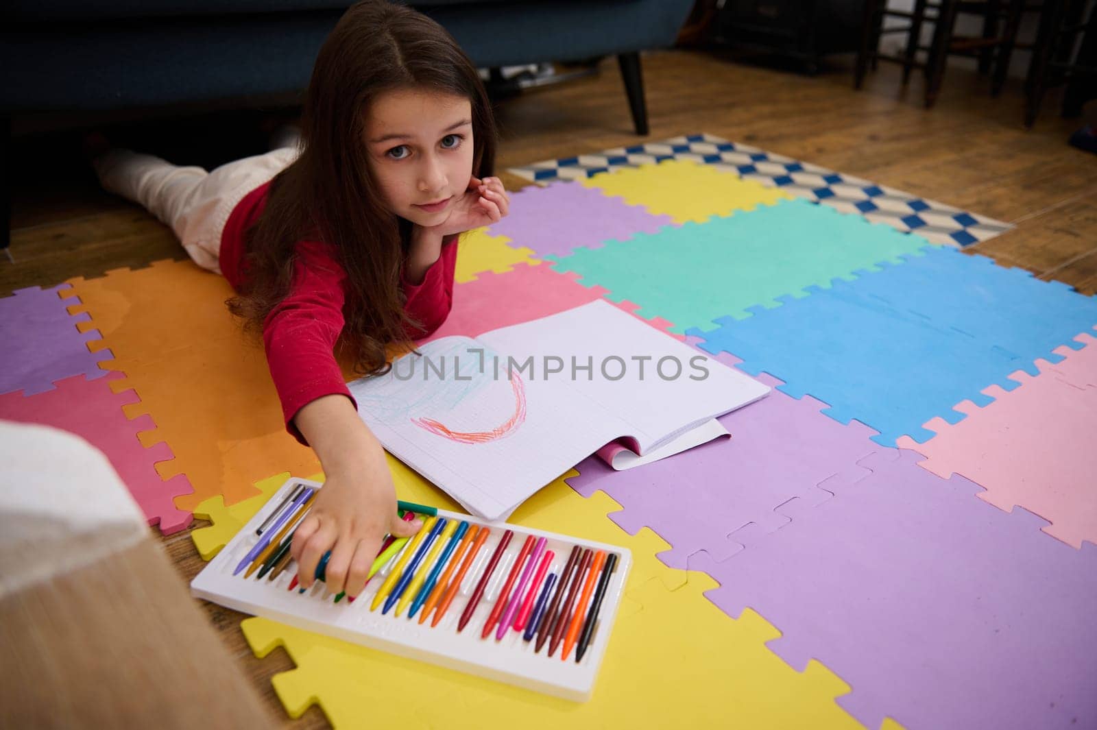 Little kid girl smiling looking at camera while taking a colorful pencil out of a box with pastel crayons, lying on her belly on a multicolored puzzle carpet. People. Education. Back to school concept