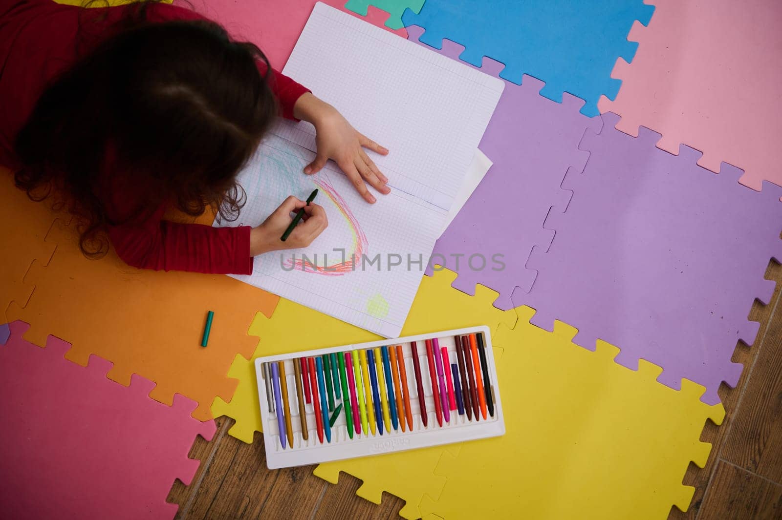 Little girl smiling drawing a picture, lying on her belly on a multicolored puzzle carpet. People. Education. Back to school concept