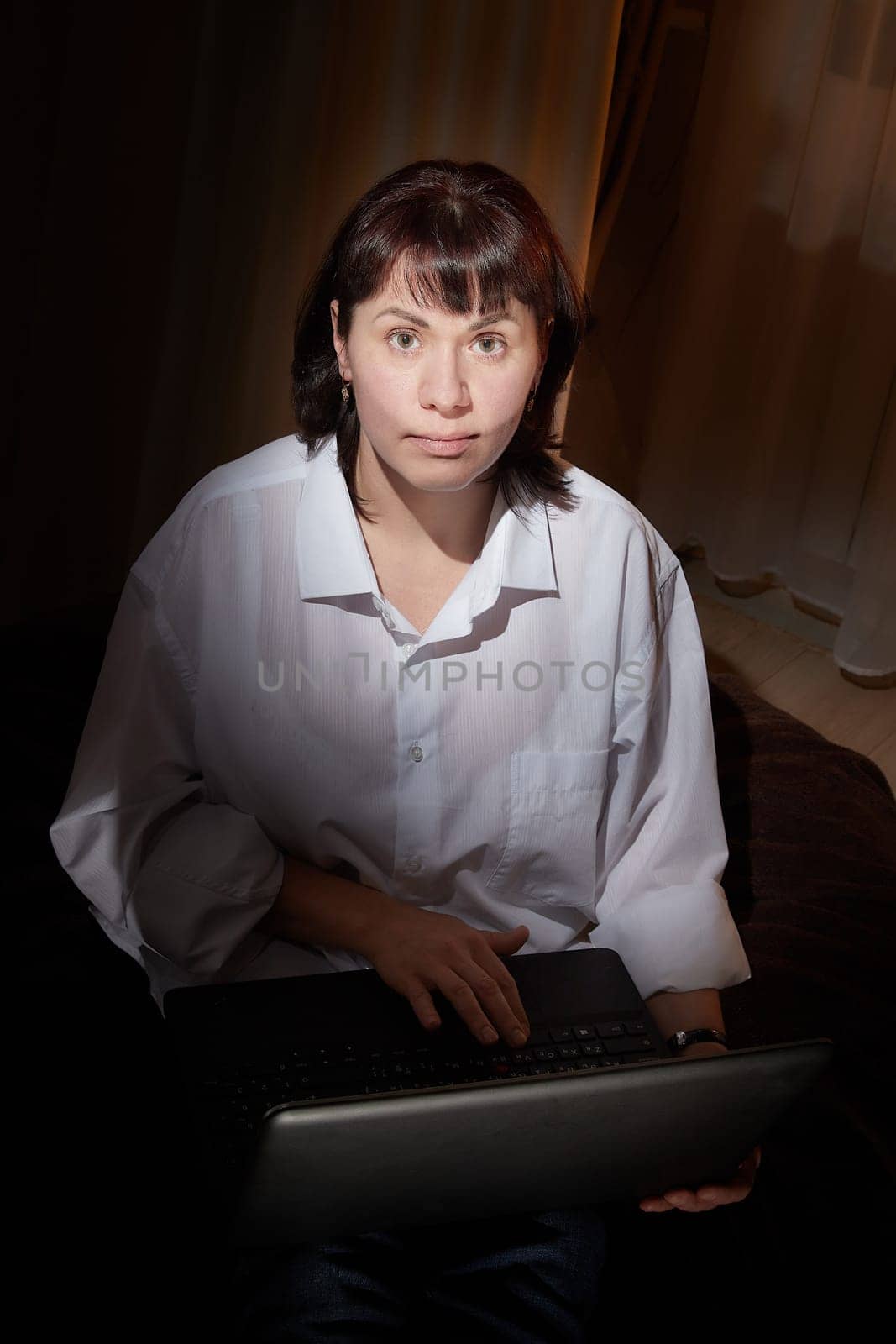 A girl in white shirt with a laptop in a dark room. A middle-aged woman works online remotely in the evening. The student is preparing for the exam at night. Blogger or freelancer in the chat