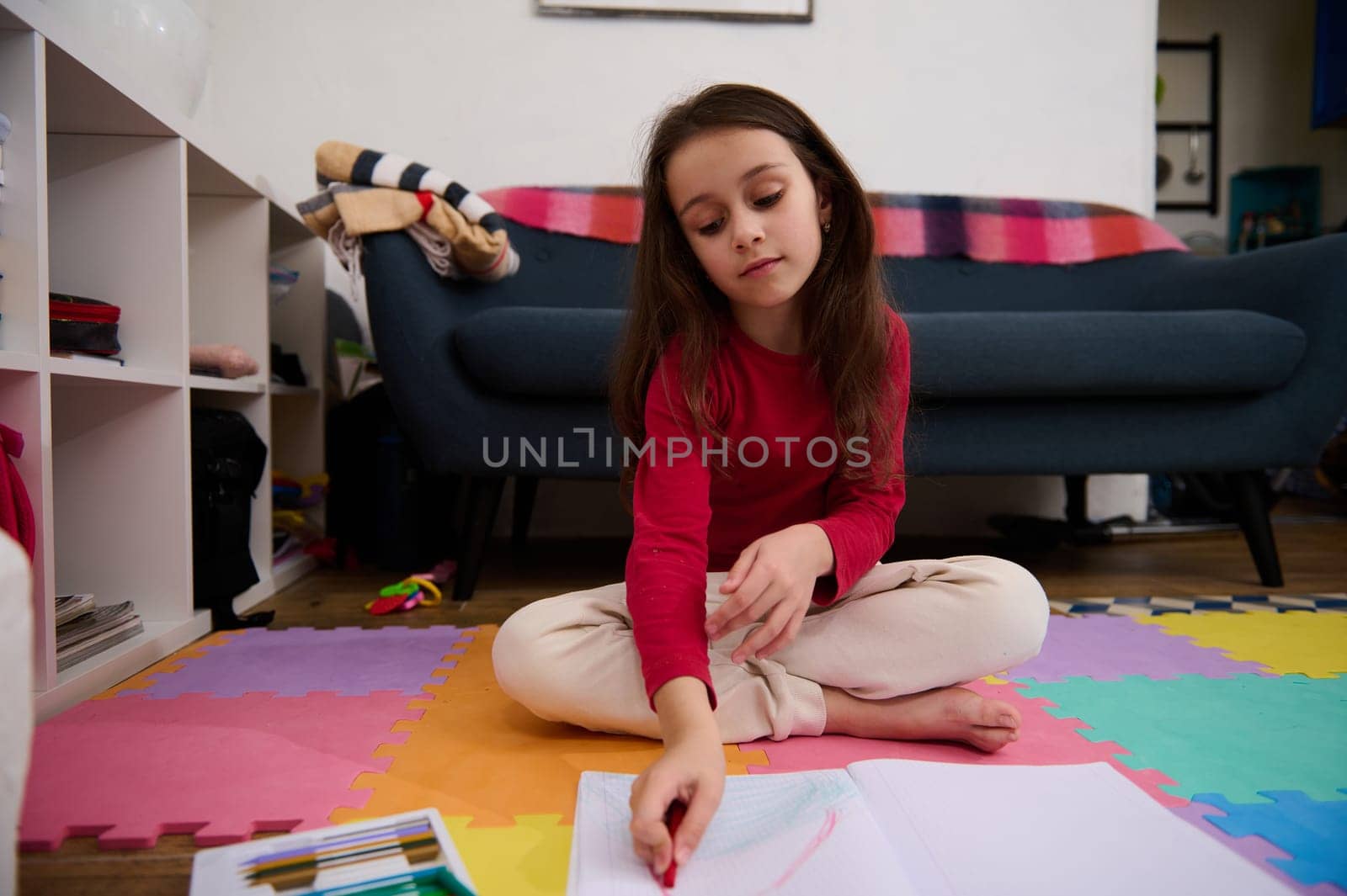 Little child girl with long hair, sitting on the puzzle carpet at home, doing homework, studying, drawing picture with colorful pencils. People, kids education and entertainment. Art and creativity by artgf