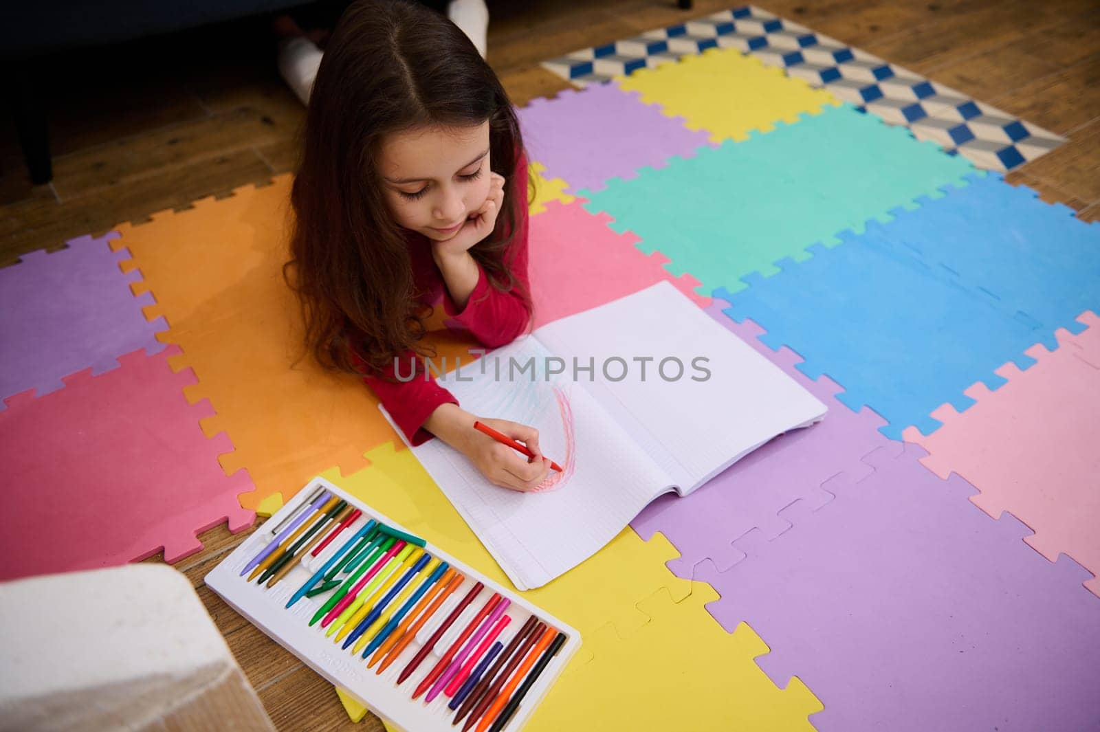 Top view of a cute little child girl lying down on a multi colored puzzle carpet at home, doing homework, drawing creative image with cloud and rainbow. Art and creativity. Kids education nd hobbies by artgf