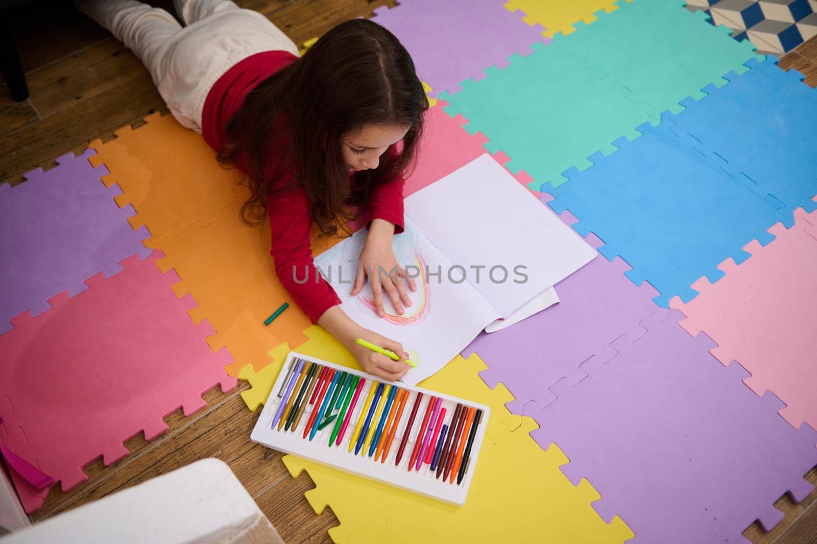 Overhead view of a talented little child, school girl taking out colorful pencil from pencil case, drawing beautiful cloud with rainbow, lying on a multi colored puzzle carpet in cozy home interior.