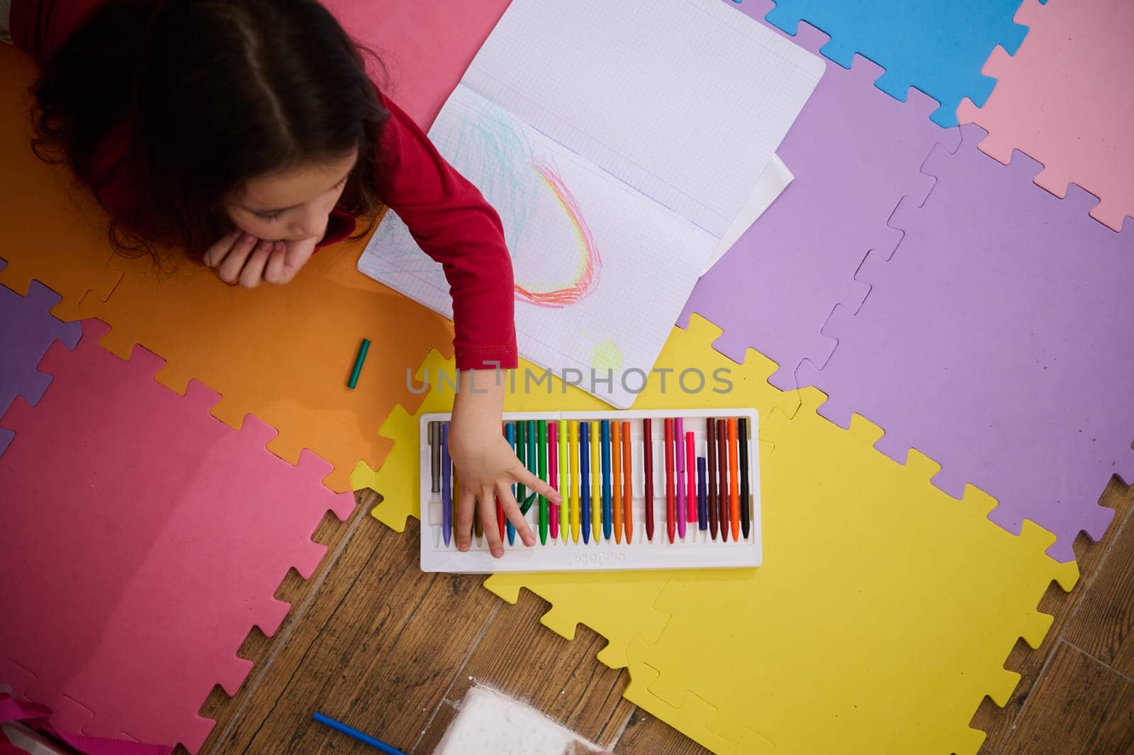 View from above of a little girl taking out colorful pencil from pencil case, drawing beautiful cloud with rainbow, lying on a multi colored puzzle carpet in cozy home interior. Kids education by artgf