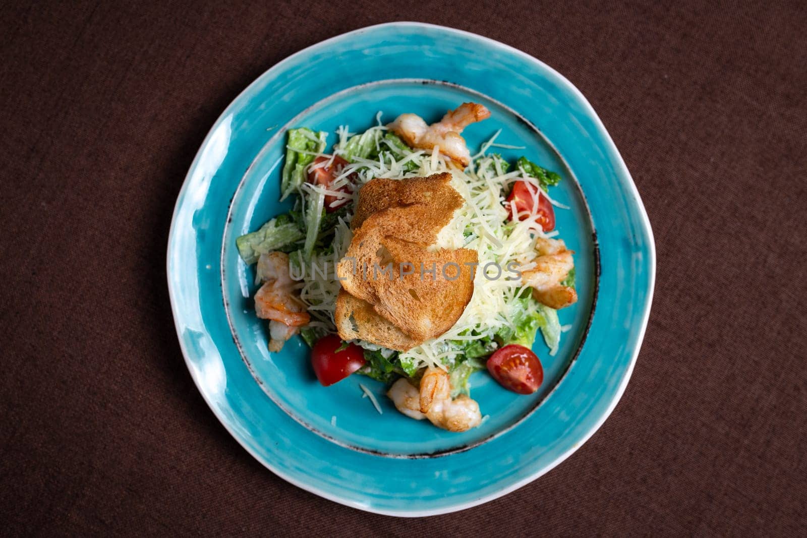 Healthy and delicious caesar salad with fresh greens, juicy tomatoes, and tasty shrimps on a blue plate. Isolated on a brown background.