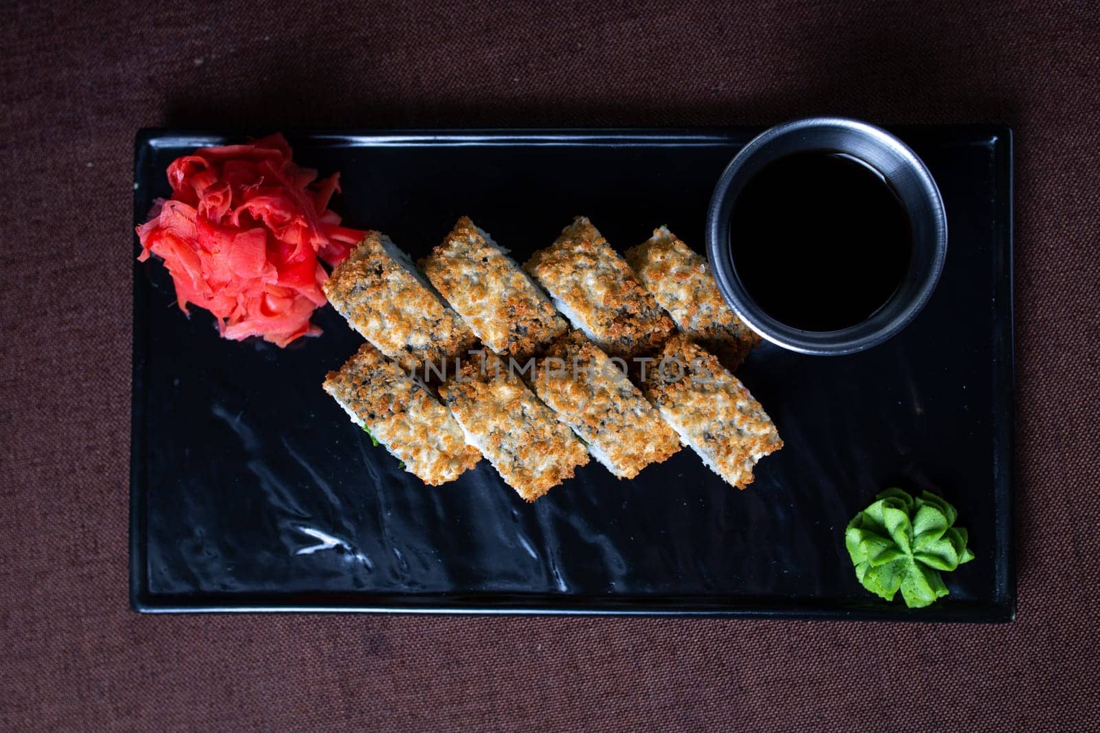 Deep-fried sushi roll with chopsticks and soy sauce on a black plate, made with rice, seaweed, fillings, and golden brown.