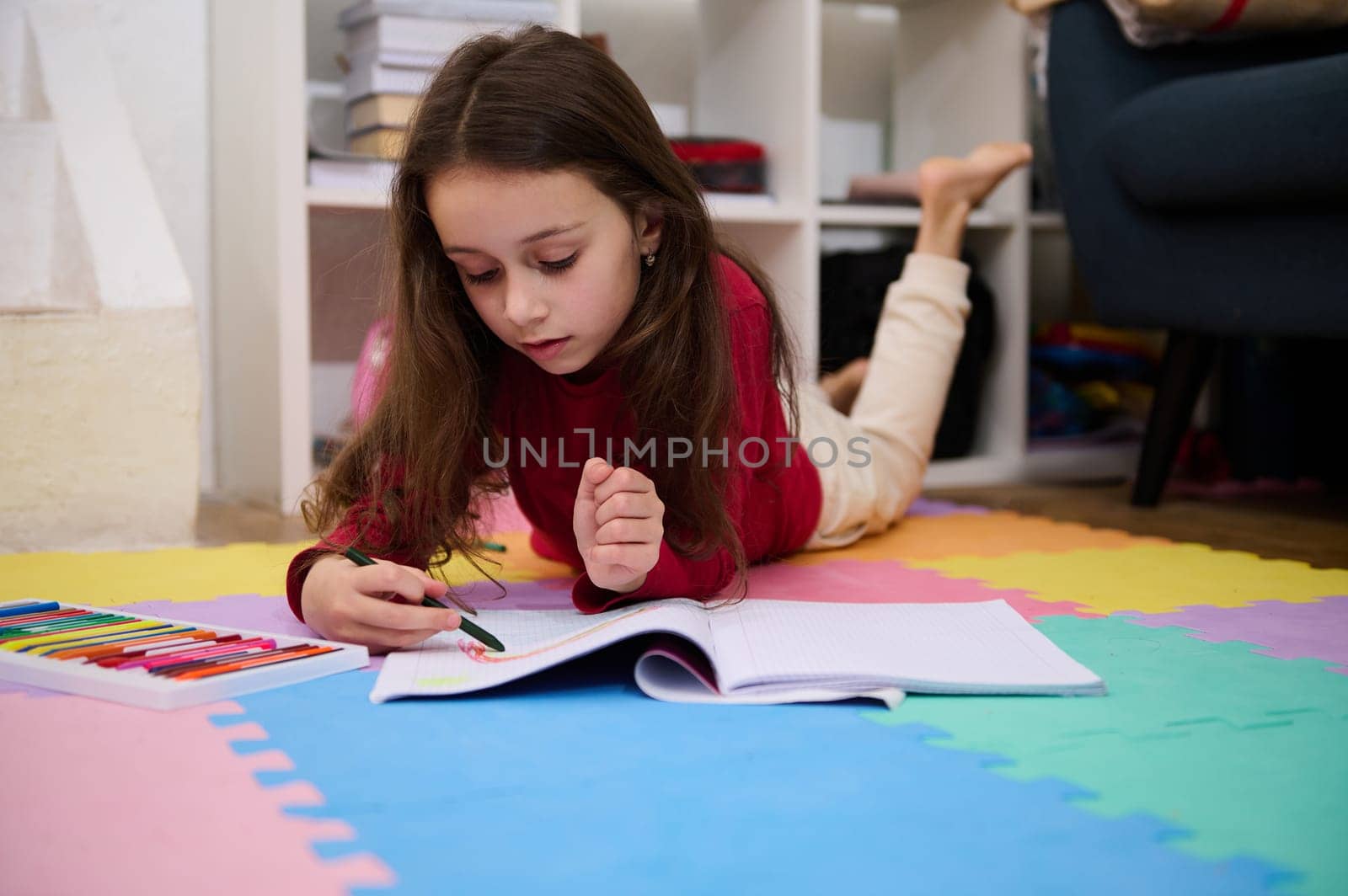 Authentic portrait of Caucasian elementary age school child girl doing homework, drawing picture with colorful, pastel pencils, writing and reading at home, lying on a multi colored puzzle carpet