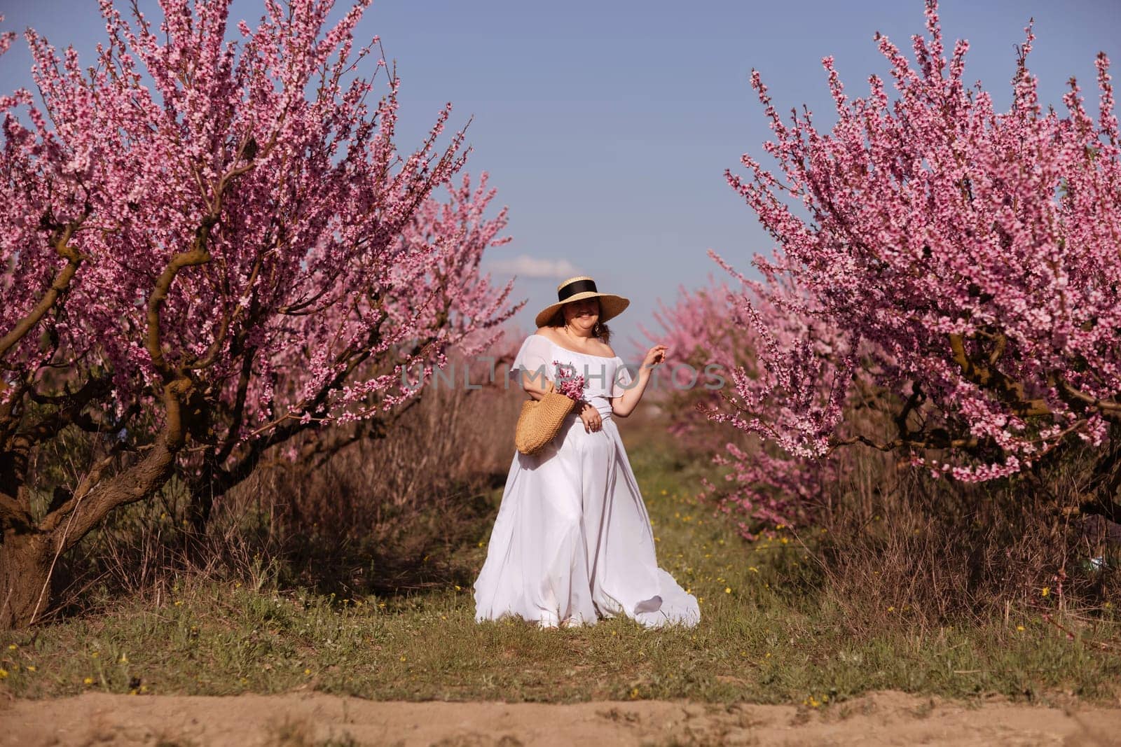 Woman blooming peach orchard. Against the backdrop of a picturesque peach orchard, a woman in a long white dress and hat enjoys a peaceful walk in the park, surrounded by the beauty of nature