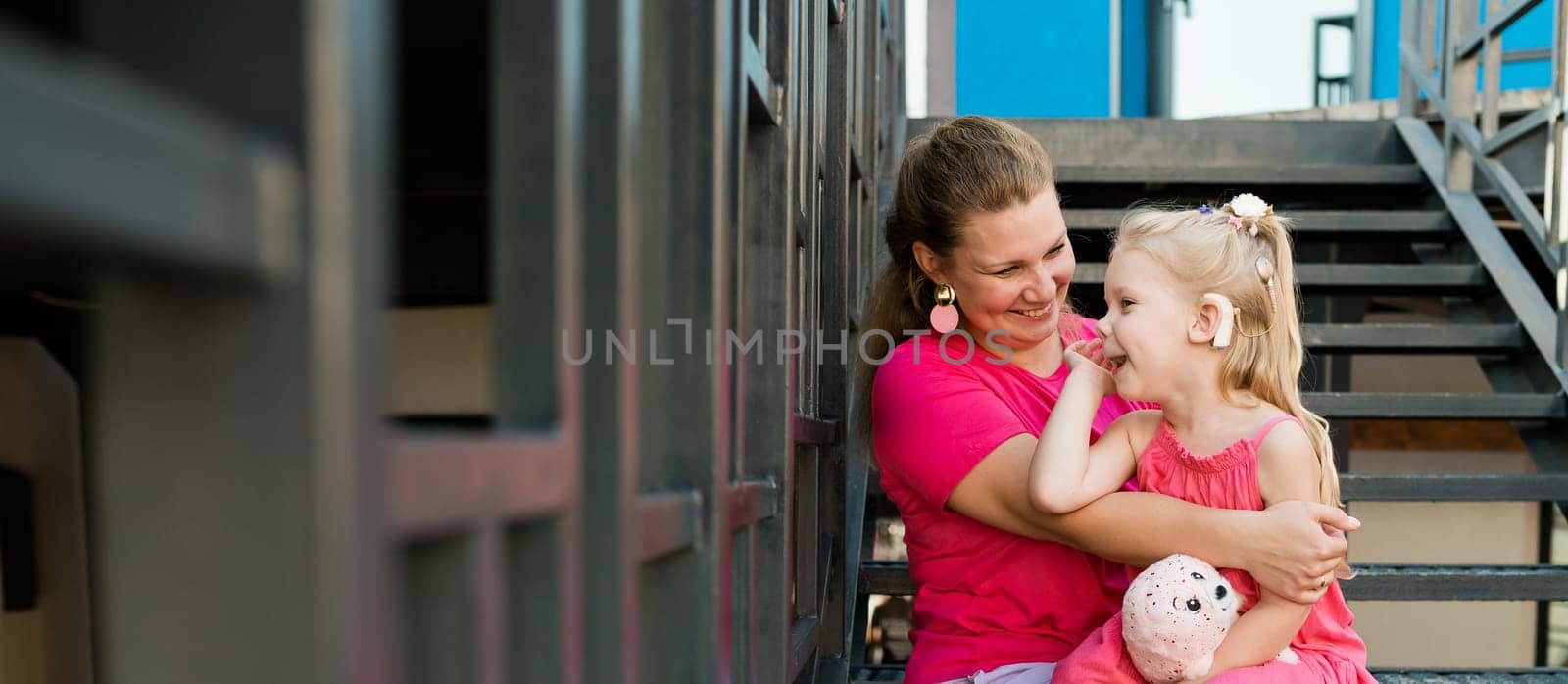 Blonde little girl with cochlear implant playing with her mother outdoor. Hear impairment deaf and health concept. Diversity and inclusion. Copy space.