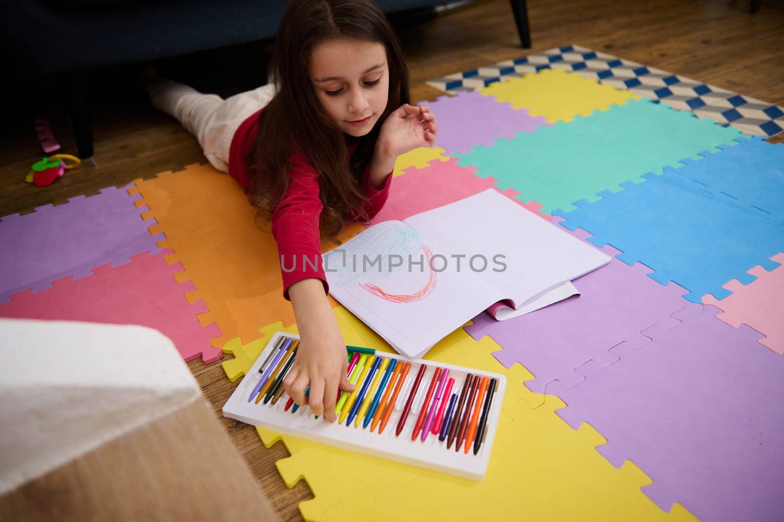 Top view of charming little girl drawing using color pastel crayons while lying on a colorful multicolored puzzle carpet on the floor in her room at home. Kids entertainment and education