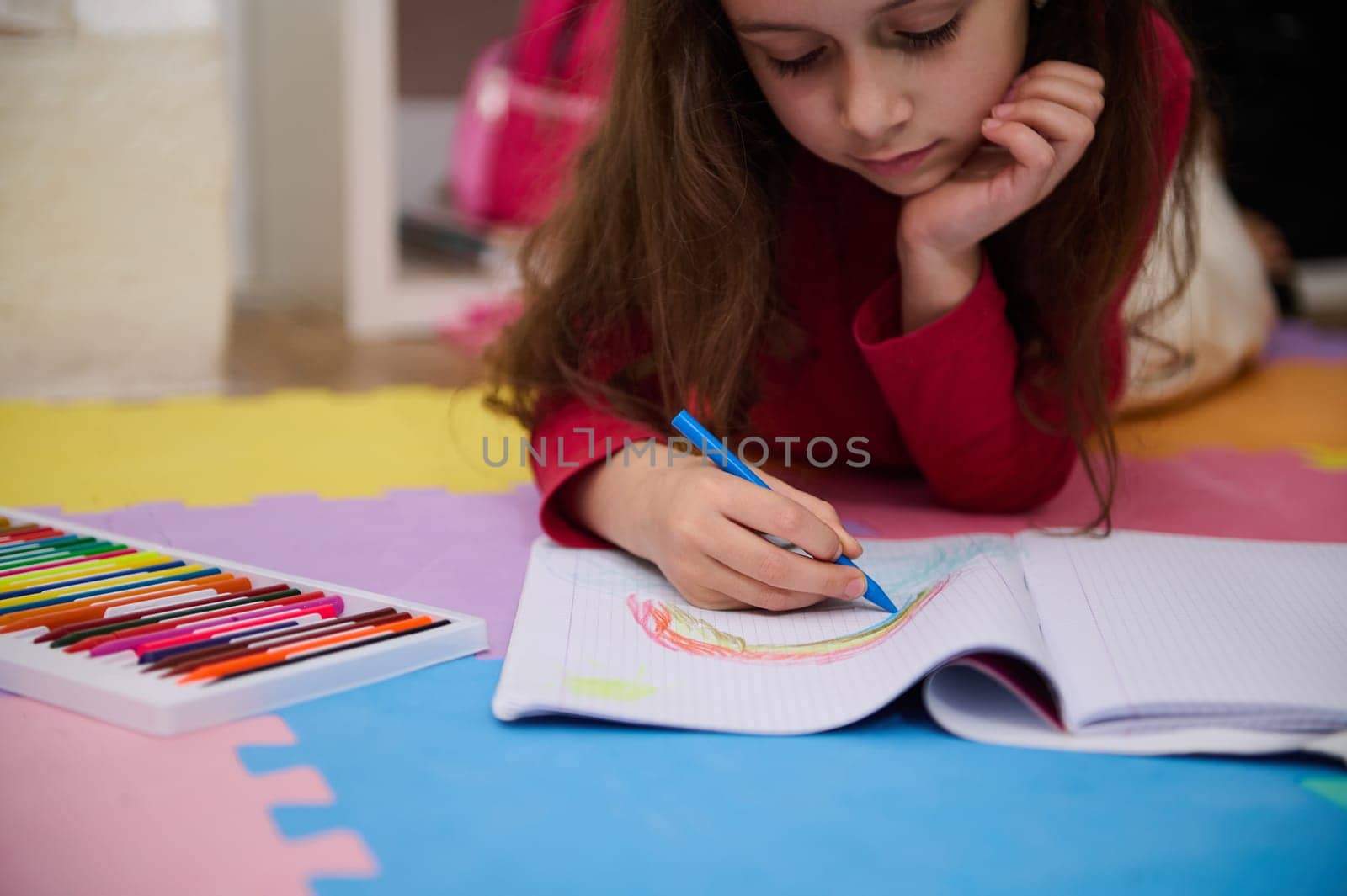 Close-up portrait of a beautiful little child girl drawing rainbow with colorful pastel pencils, lying on a multicolored puzzle carpet in her room at home. Art and creativity concept. Kids development