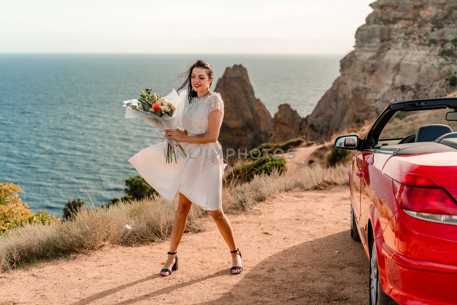 A woman in a white dress is standing next to a red convertible car. She is holding a bouquet of flowers and she is in a happy mood. The scene suggests a romantic or special occasion
