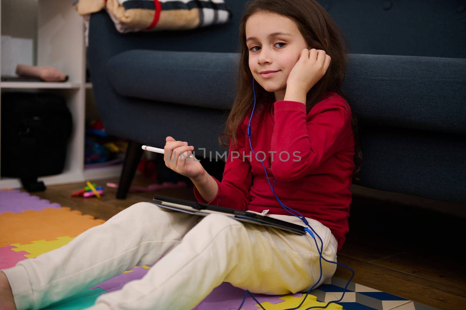 Portrait of a lovely child girl using digital tablet, studying online from home, smiling looking at camera, sitting on colorful puzzle carpet. Kids education. Distance learning. Autism. Diversity by artgf