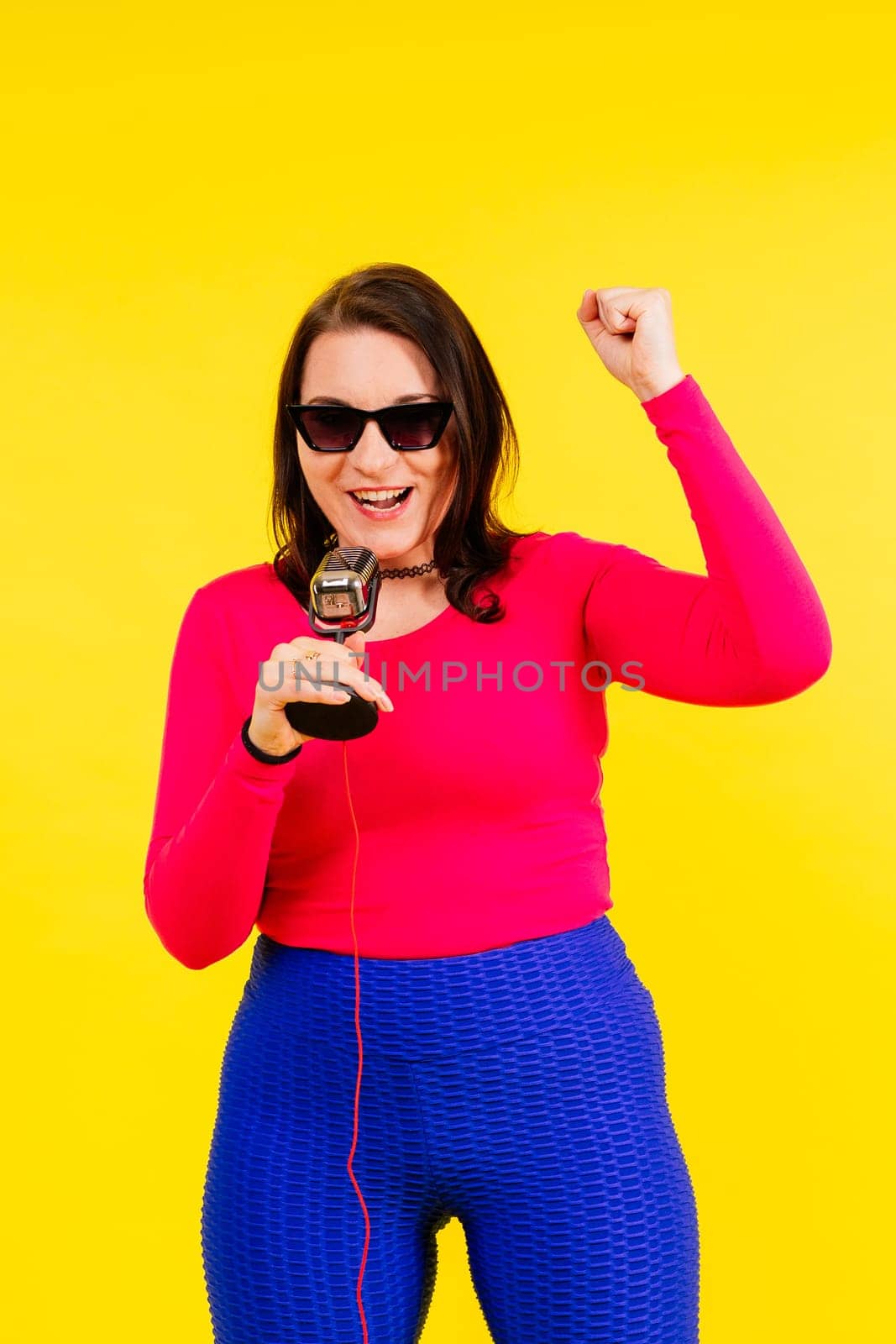 Woman in sport clothes sings into wired microphone and claps her hands on a yellow background