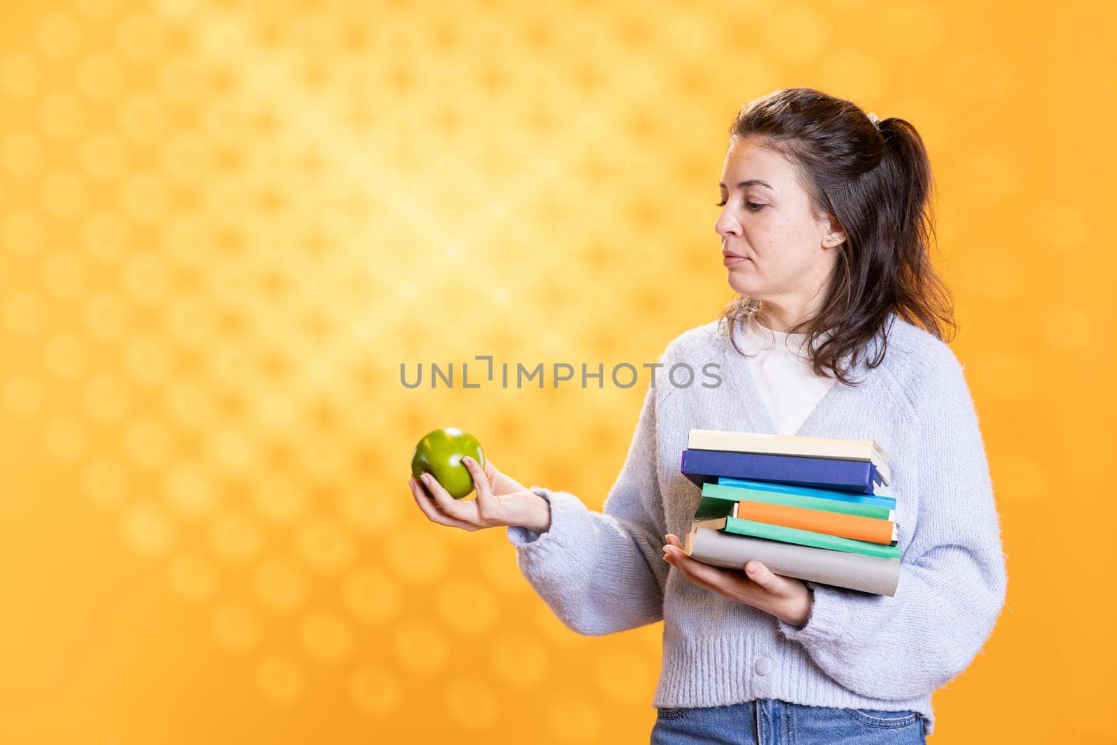 Woman with stack of books in hands enjoying fresh apple, living healthy, isolated over studio background. Bookworm holding pile of novels and green fruit, enjoying bio snack