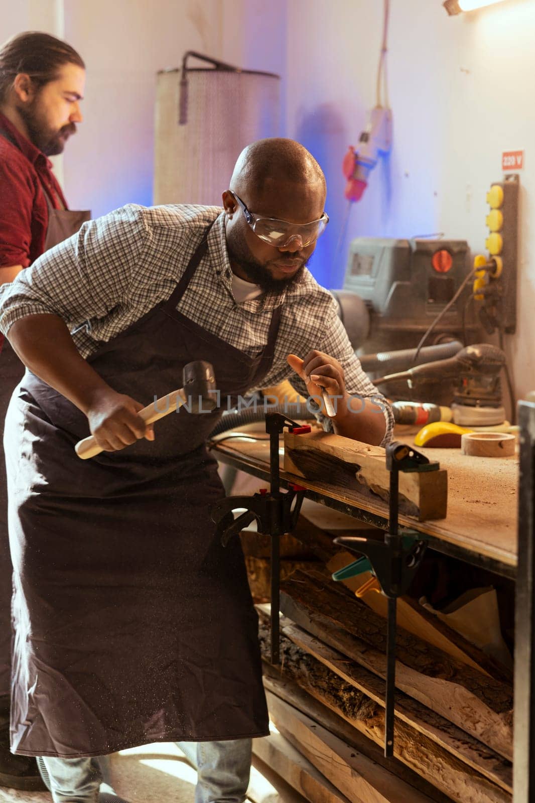 Craftsperson carving designs into wood using chisel and hammer next to coworker by DCStudio