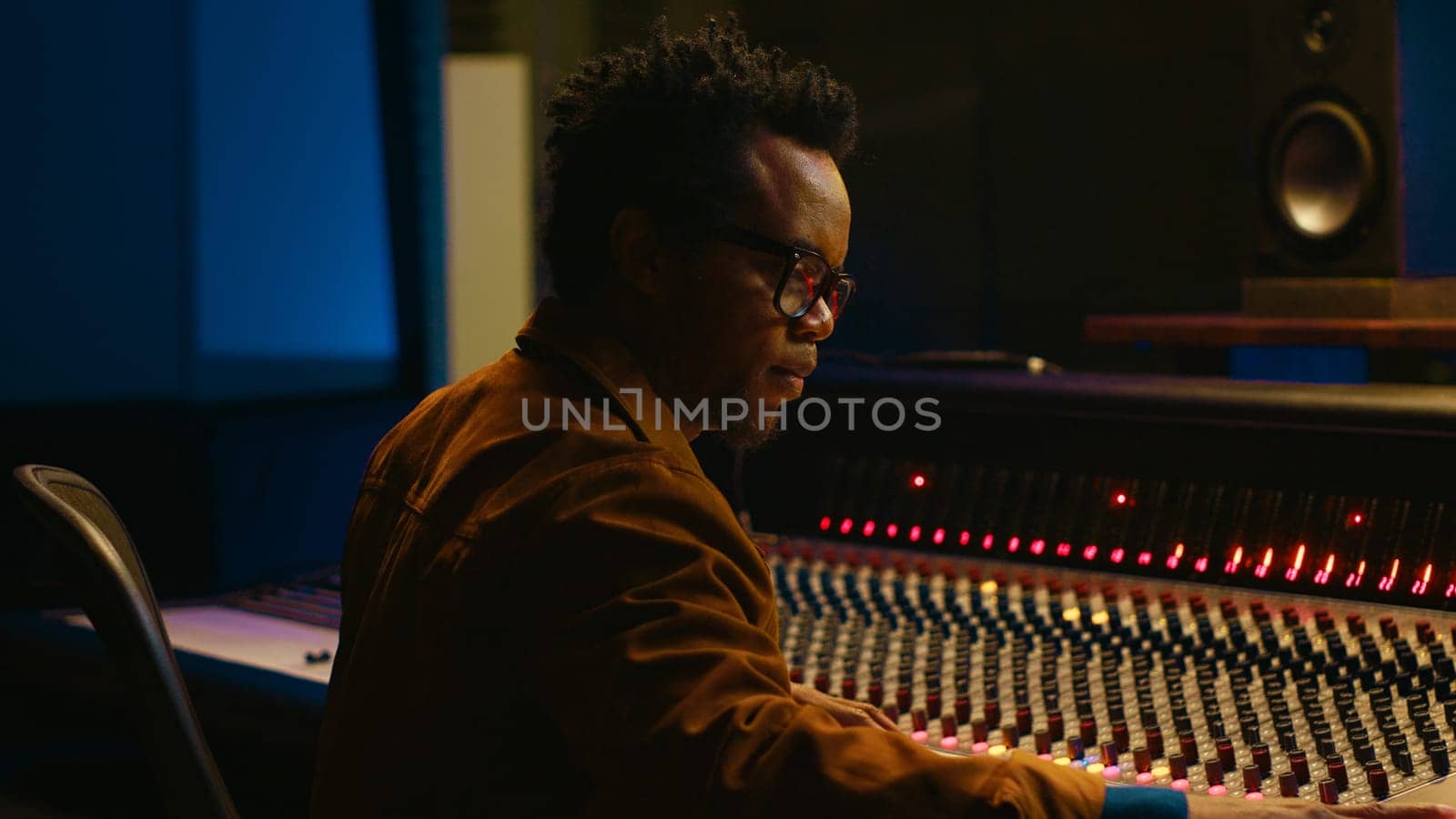 Portrait of african american audio technician uses mixing console with knobs and sliders in control room, editing recordings and adding sound effects. Skilled music producer at mixer. Camera A.